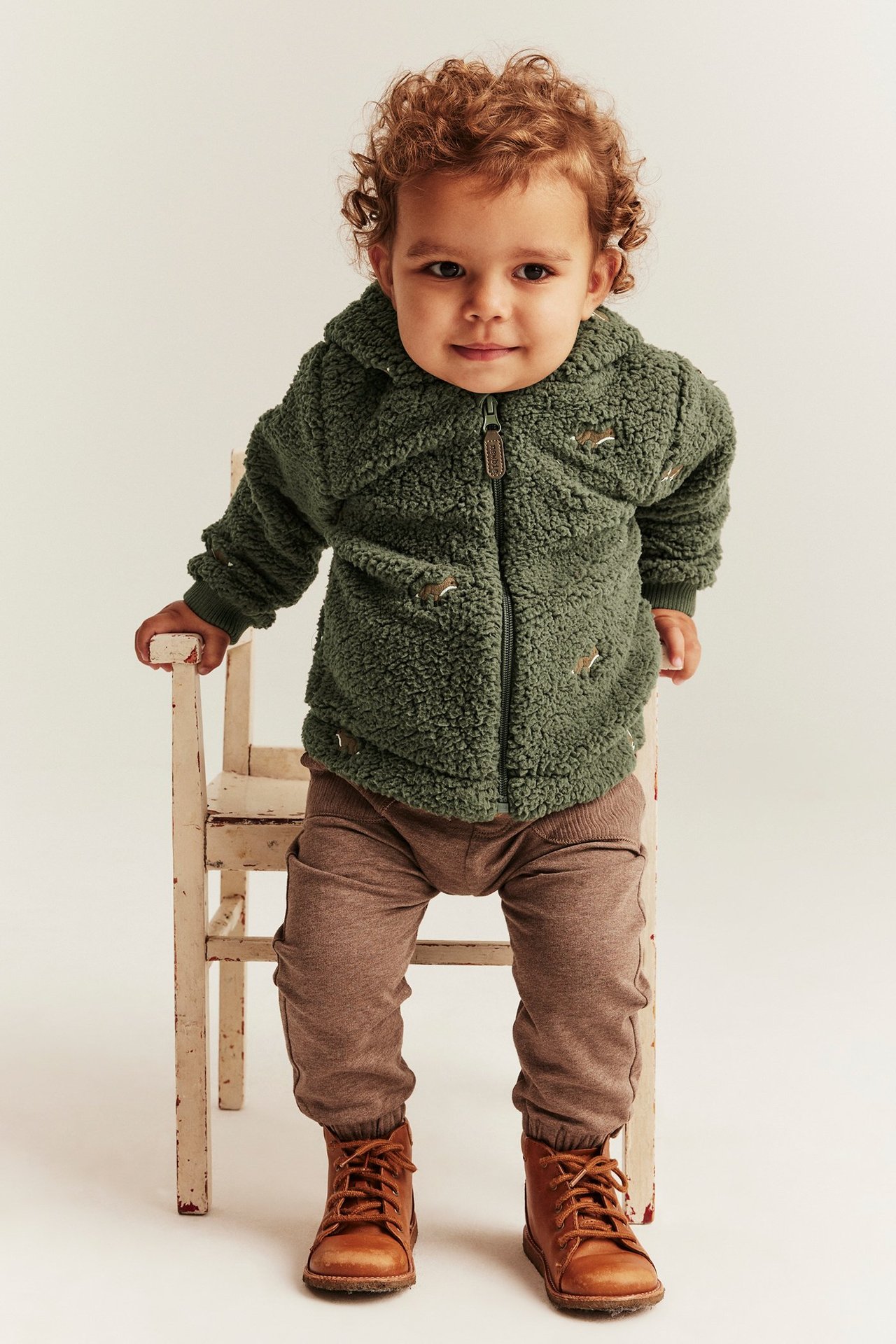 Vorderansicht eines Babys in dunkelgrüner Teddyjacke mit Fuchsstickerei, brauner Hose und braunen Schnürstiefeln.