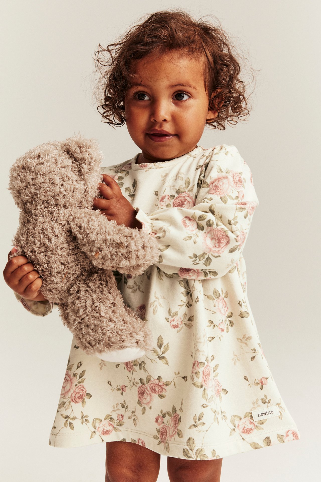 Front view: Kid in a light beige floral jersey dress with puff sleeves, holding a brown teddy bear.