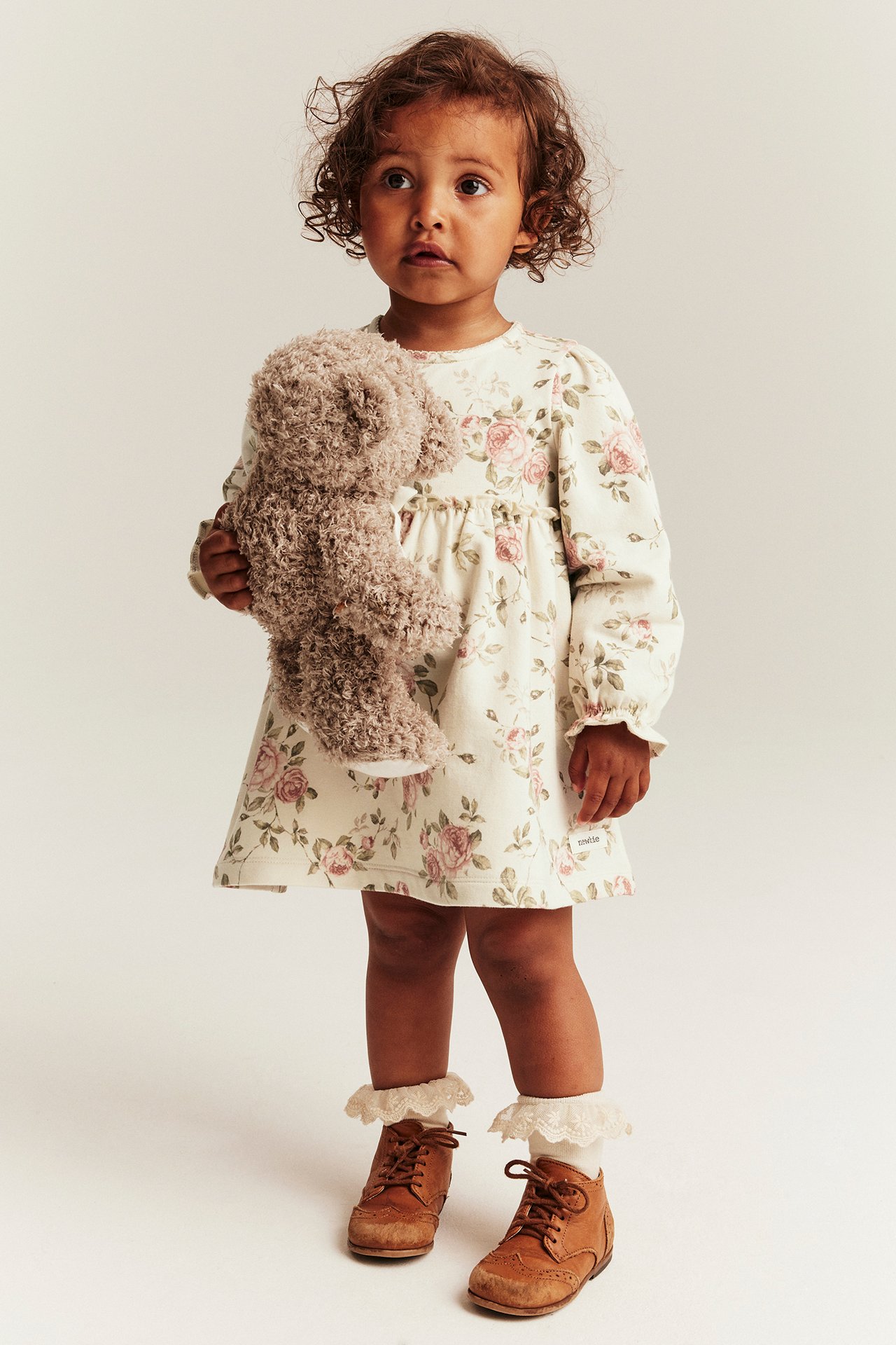 Front view: Baby in light beige floral jersey dress, holding a brown teddy bear, white lace socks, brown boots.