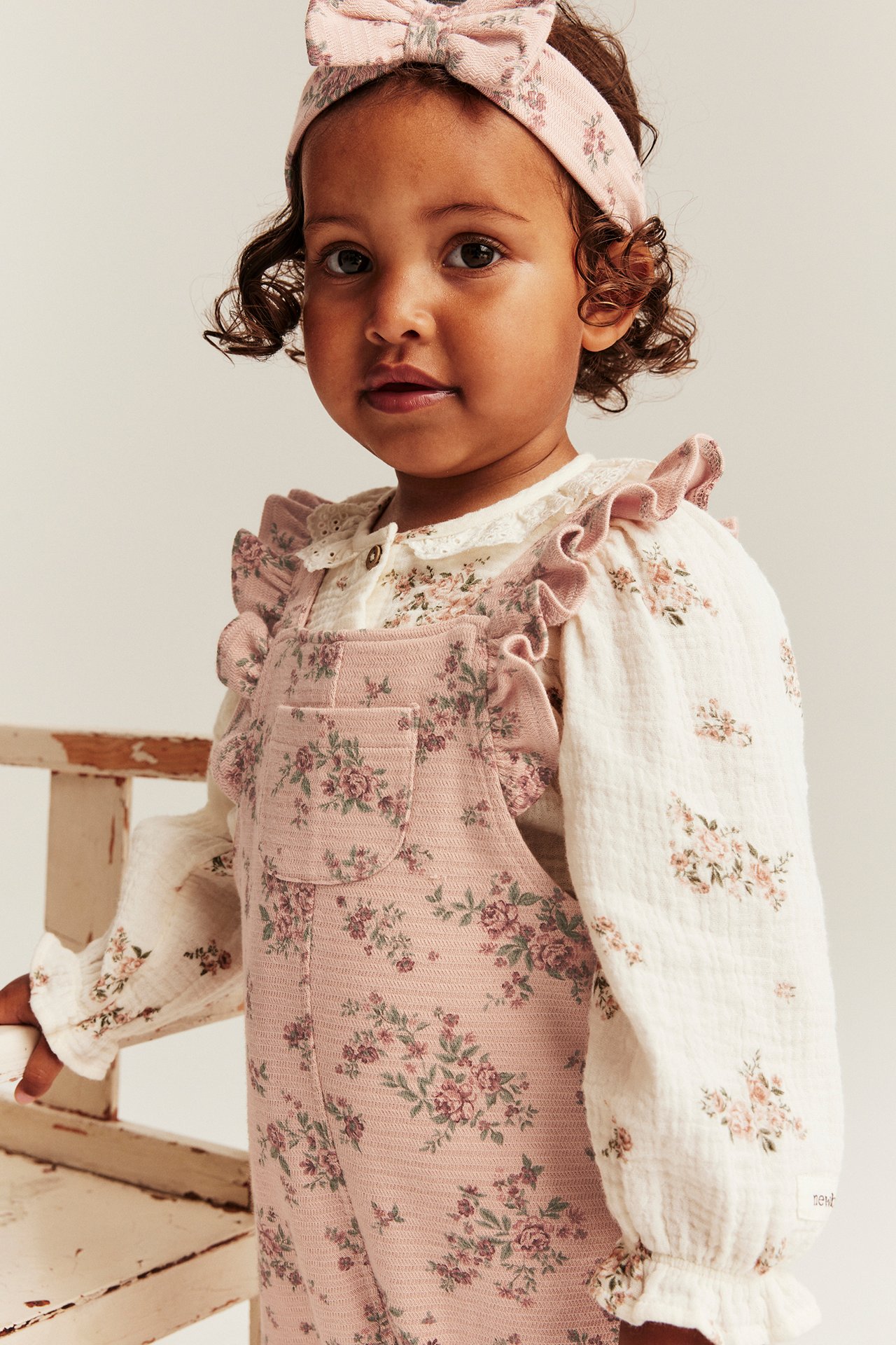 Vorderansicht: Baby in rosa geblümter Jersey-Latzhose mit Rüschen, weißem Blümchen-Oberteil und passendem Haarband.