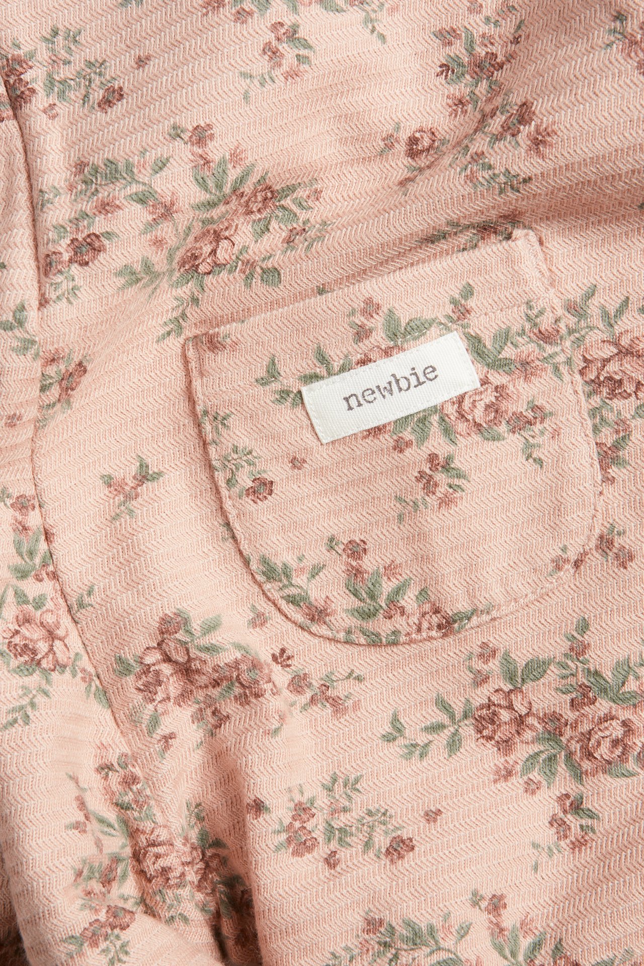 Nahaufnahme rosa geblümter Baby-Latzhose aus Strukturjersey, mit Tasche und 'newbie'-Etikett.