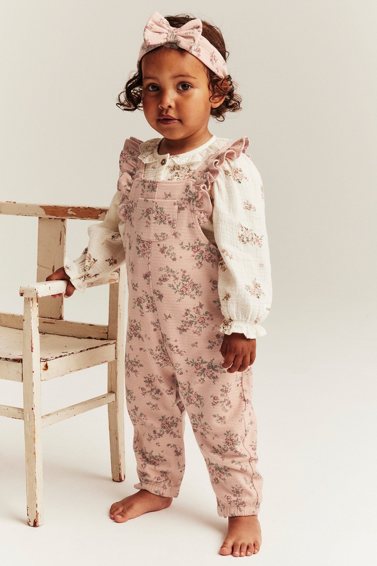 Vorderansicht: Kind in rosa geblümter Jersey-Latzhose mit Rüschen, weißer Bluse und passendem Haarband.