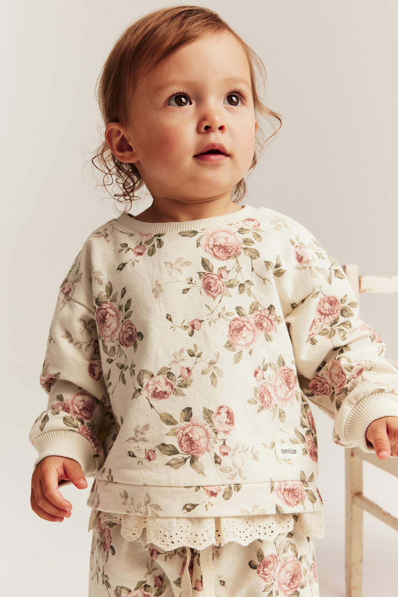Front view of a baby wearing a light beige floral sweatshirt with pink roses and matching pants with lace trim.
