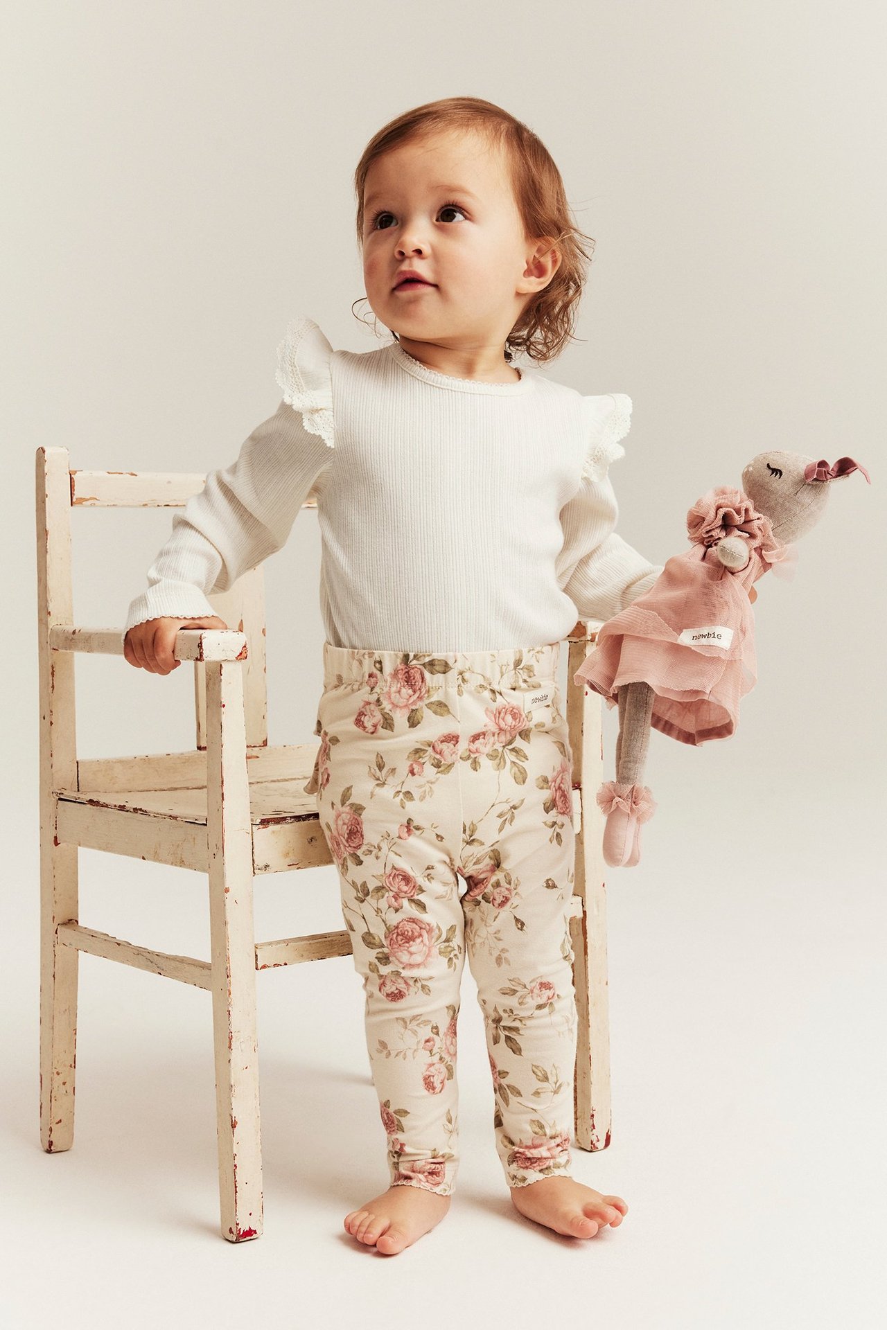 Vorderansicht: Baby trägt einen cremefarbenen Ripp-Body mit Spitzenrüschen, geblümte Leggings, hält eine Puppe.