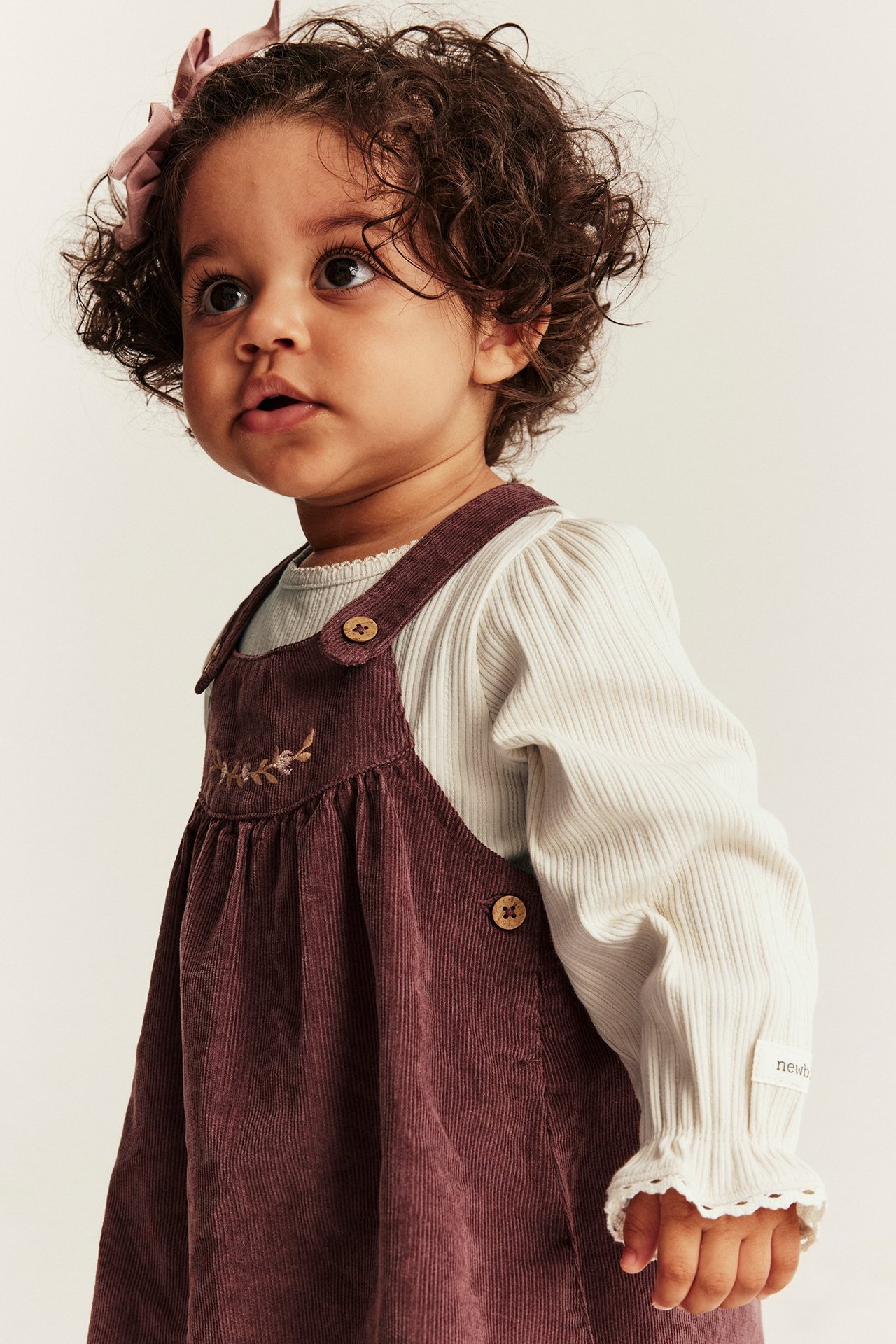 Vorderansicht: Baby in hellbeigem Rippenbody mit Spitzenbündchen, pflaumenfarbenem Cordkleid, rosa Schleife.