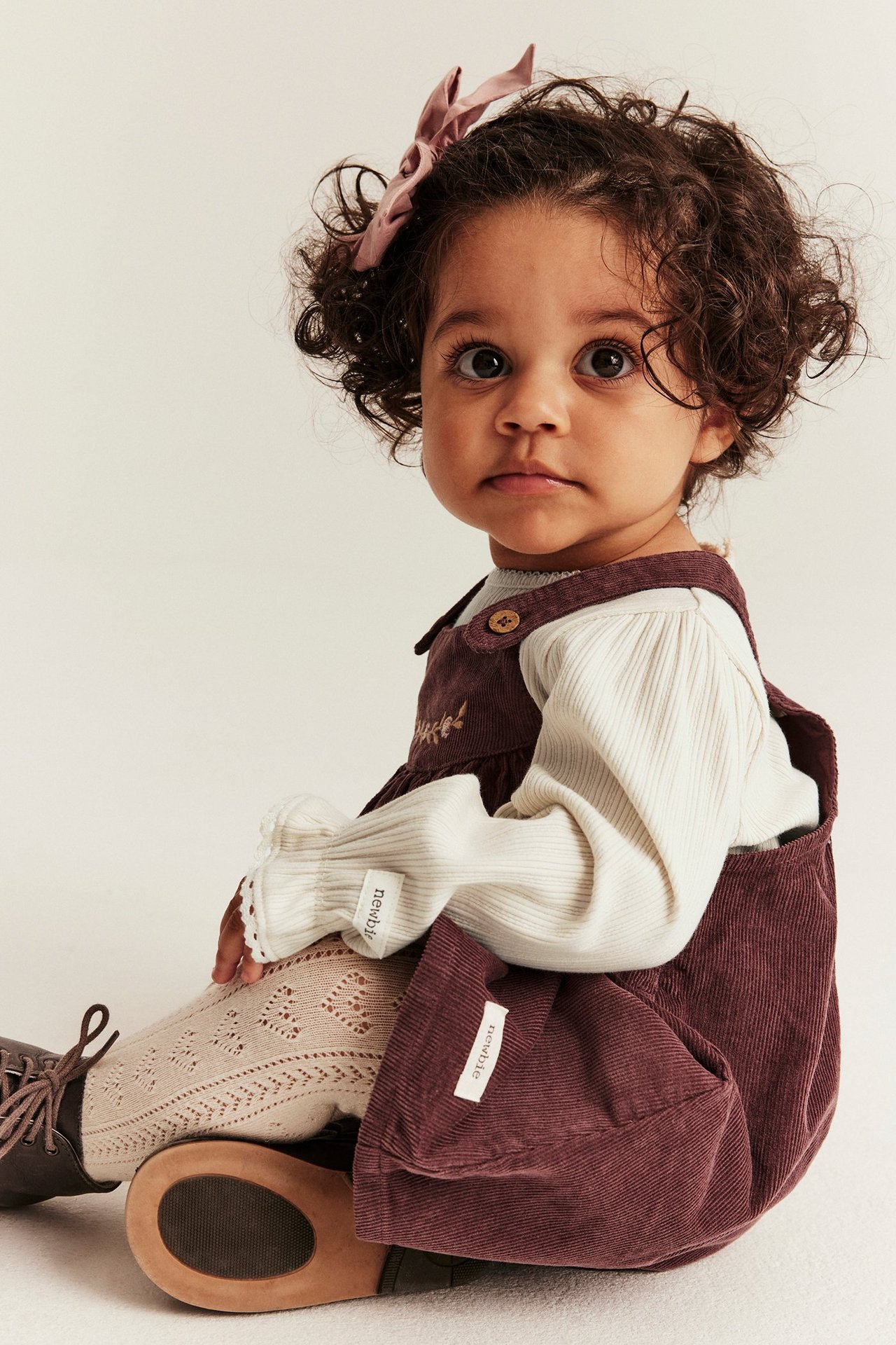 Baby trägt hellbeigen Rippen-Body mit Spitzenbündchen, dunkelroten Cord-Latzrock und gemusterte Strumpfhosen.