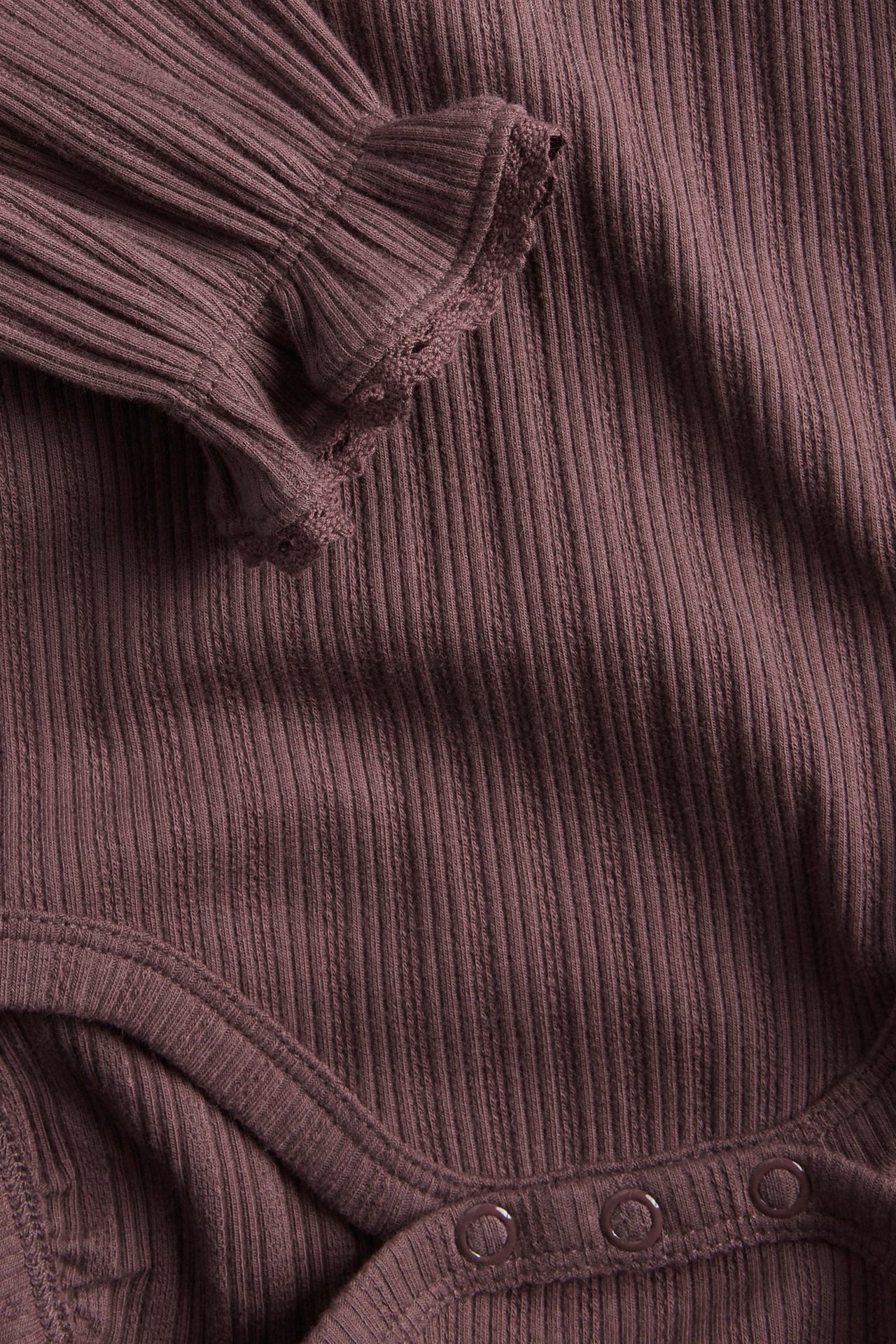 Nahaufnahme eines lila gerippten Baby-Bodys mit Rüschen und Spitzenbündchen, Druckknöpfe.