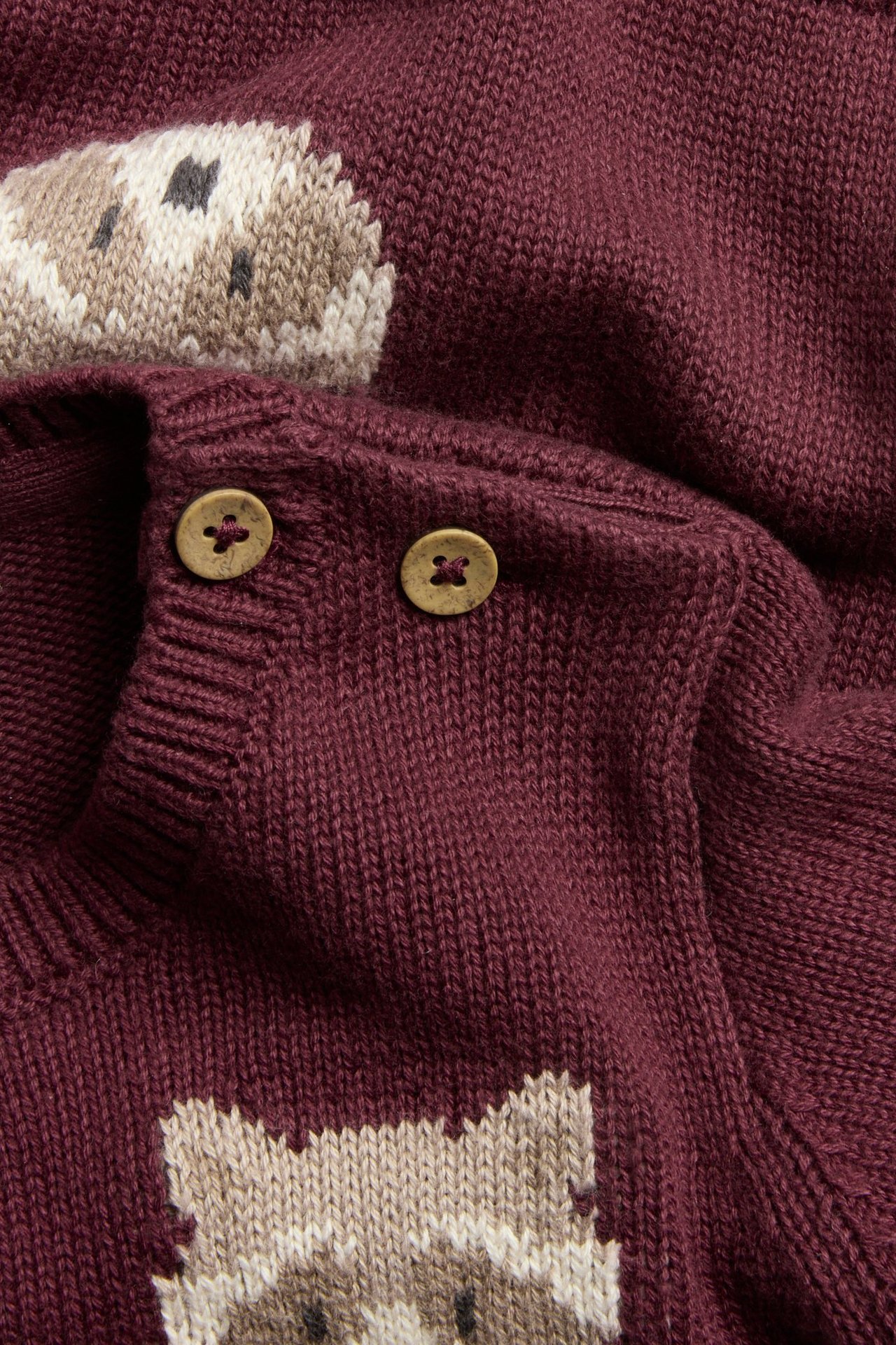 Nahaufnahme eines weinroten Jacquard-Strickpullovers für Kinder mit Waschbärmuster und zwei Holzknöpfen an der Schulter.