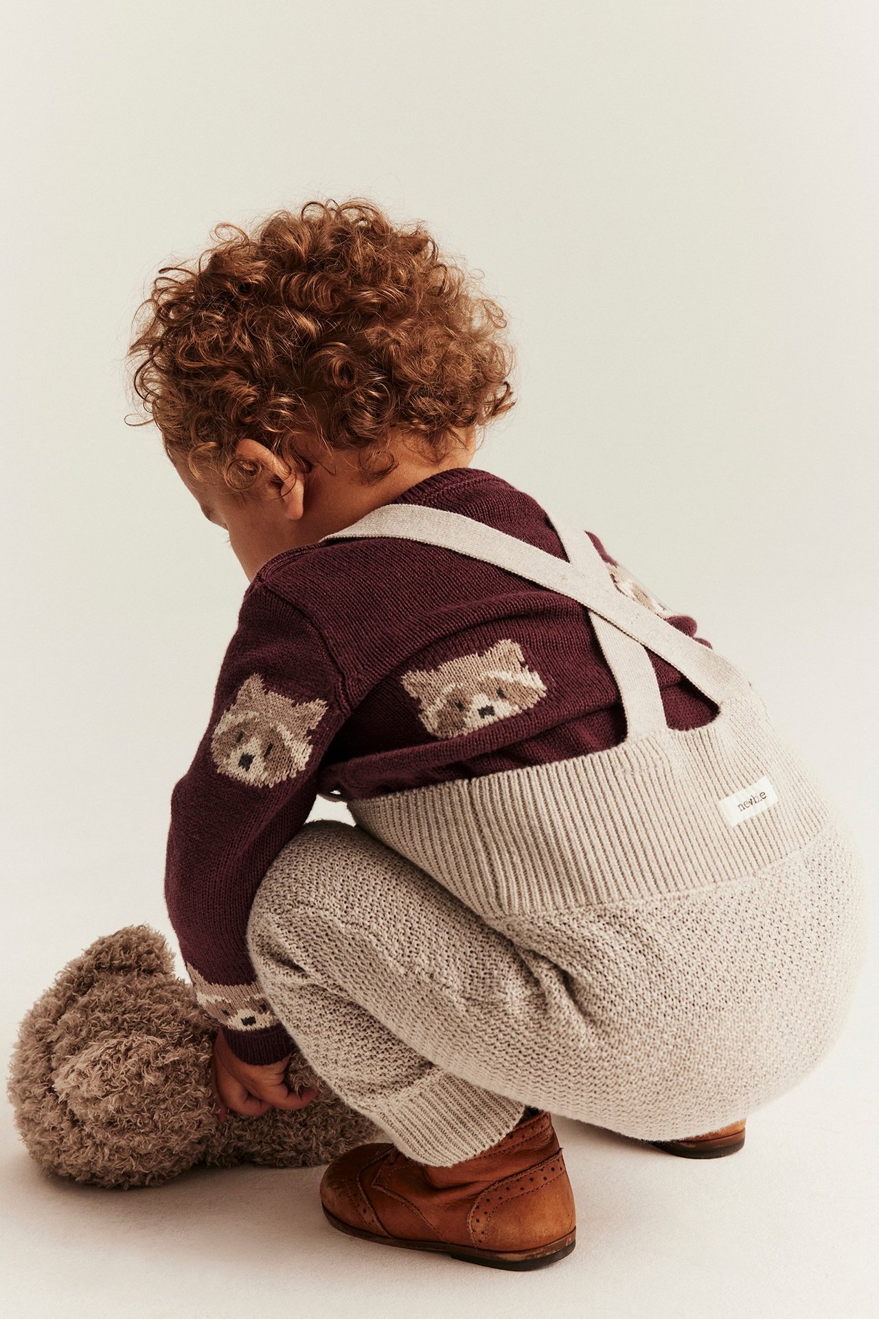 Rückenansicht: Baby in weinrotem Strickpullover mit Waschbären, hellbeiger Latzhose, braunen Schuhen, hält einen Teddybär.