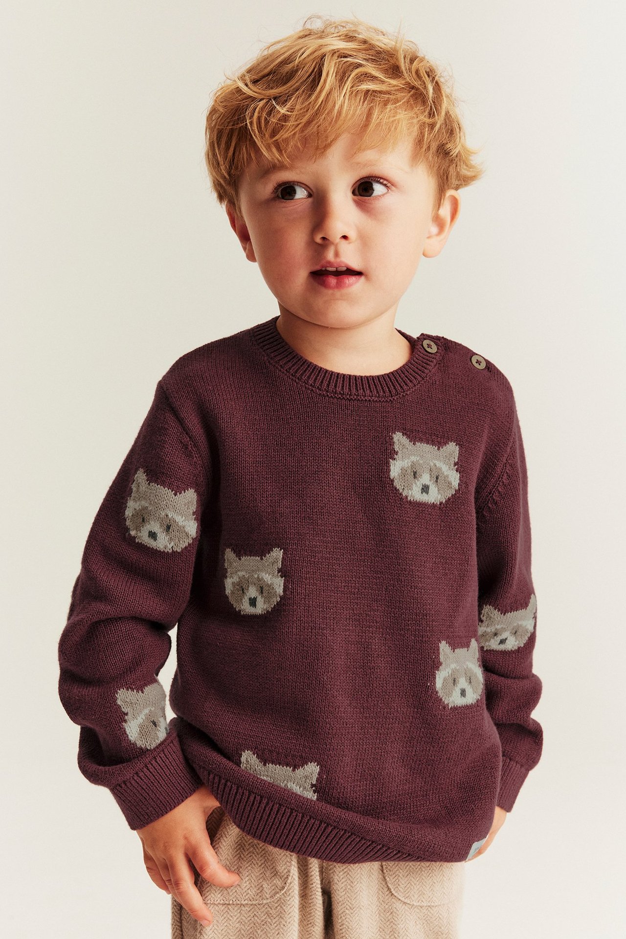 Vorderansicht: Kind in weinrotem Strickpullover aus Baumwolle mit Waschbärmuster, Schulterknöpfen und beiger Hose.