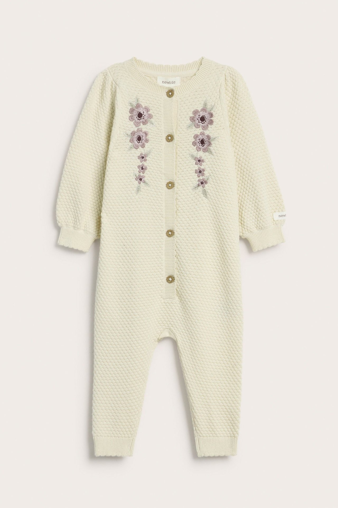 Vorderansicht eines hellbeigen strukturstrick Baby-Overalls mit lila Blumenstickerei und Knöpfen vorne.