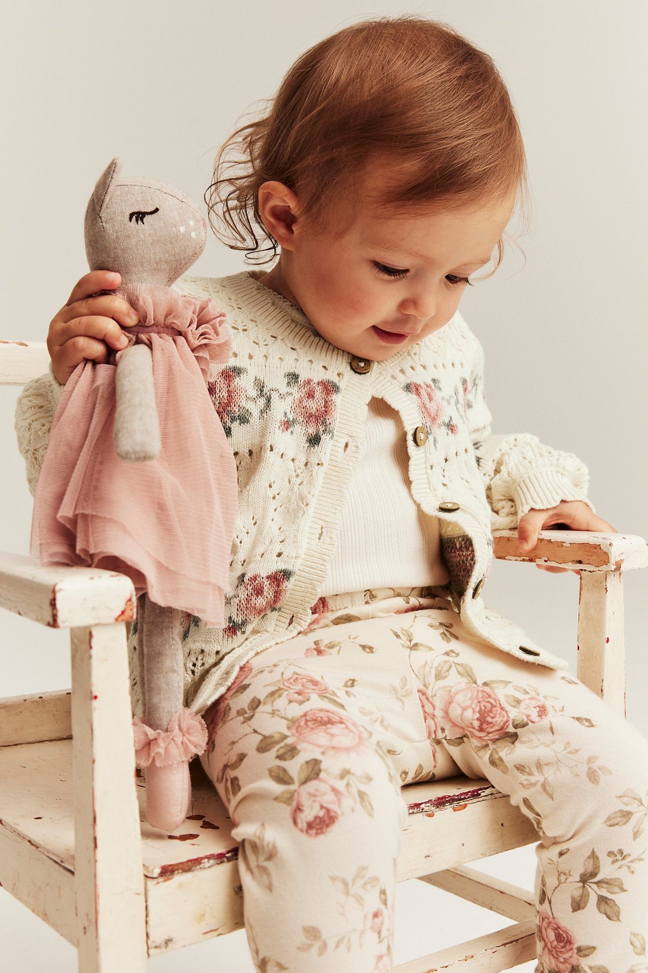 Baby in cremefarbener Jacquard-Strickjacke mit rosa Rosen, weißem Top, Blumen-Leggings, hält eine Puppe.