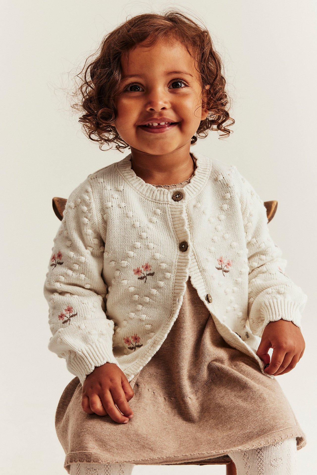 Vorderansicht eines Kindes in hellbeigem Wollmix-Cardigan mit Strickpunkten und rosa Blumenstickerei.