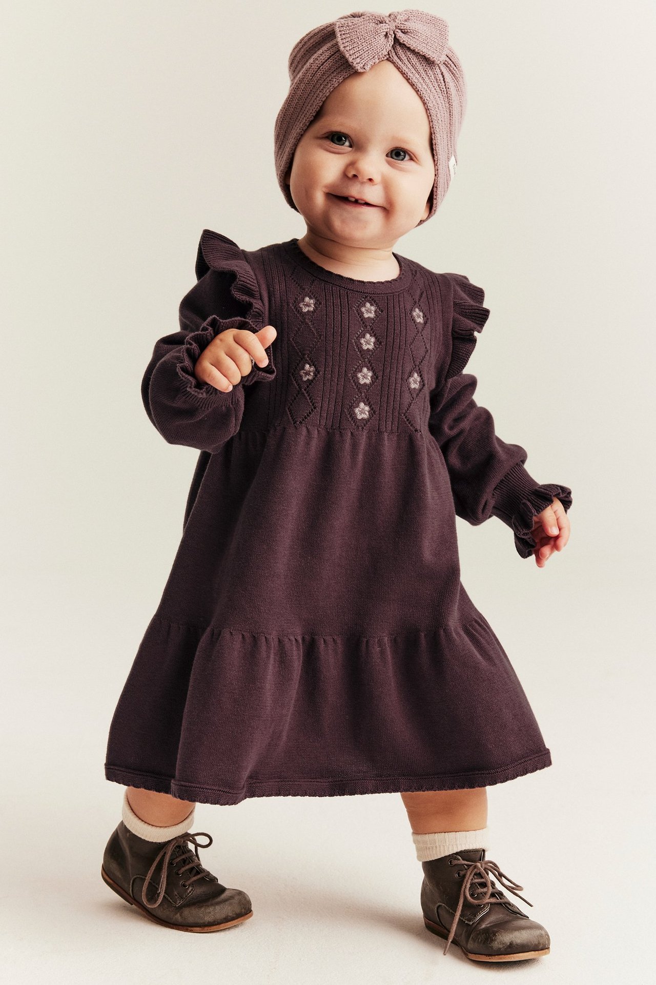 Vorderansicht: Baby in dunkel-lila Strickkleid mit Rüschen, Stickerei, rosa Turban und braunen Stiefeln.