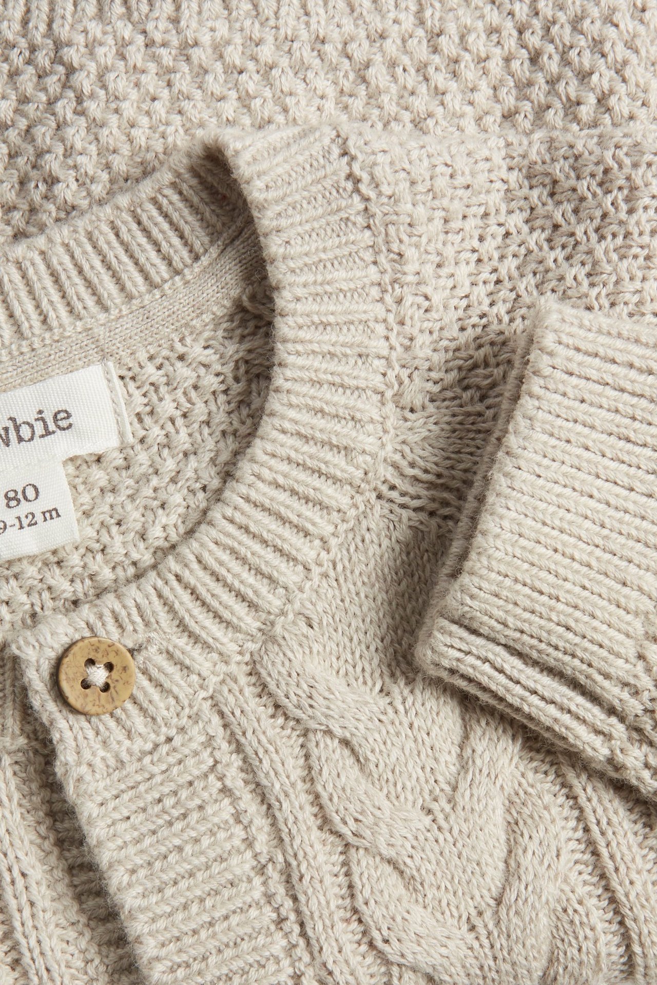 Nahaufnahme einer beigefarbenen melierten strukturgestrickten Baby-Strickjacke mit Holzknopf und Rippdetails.