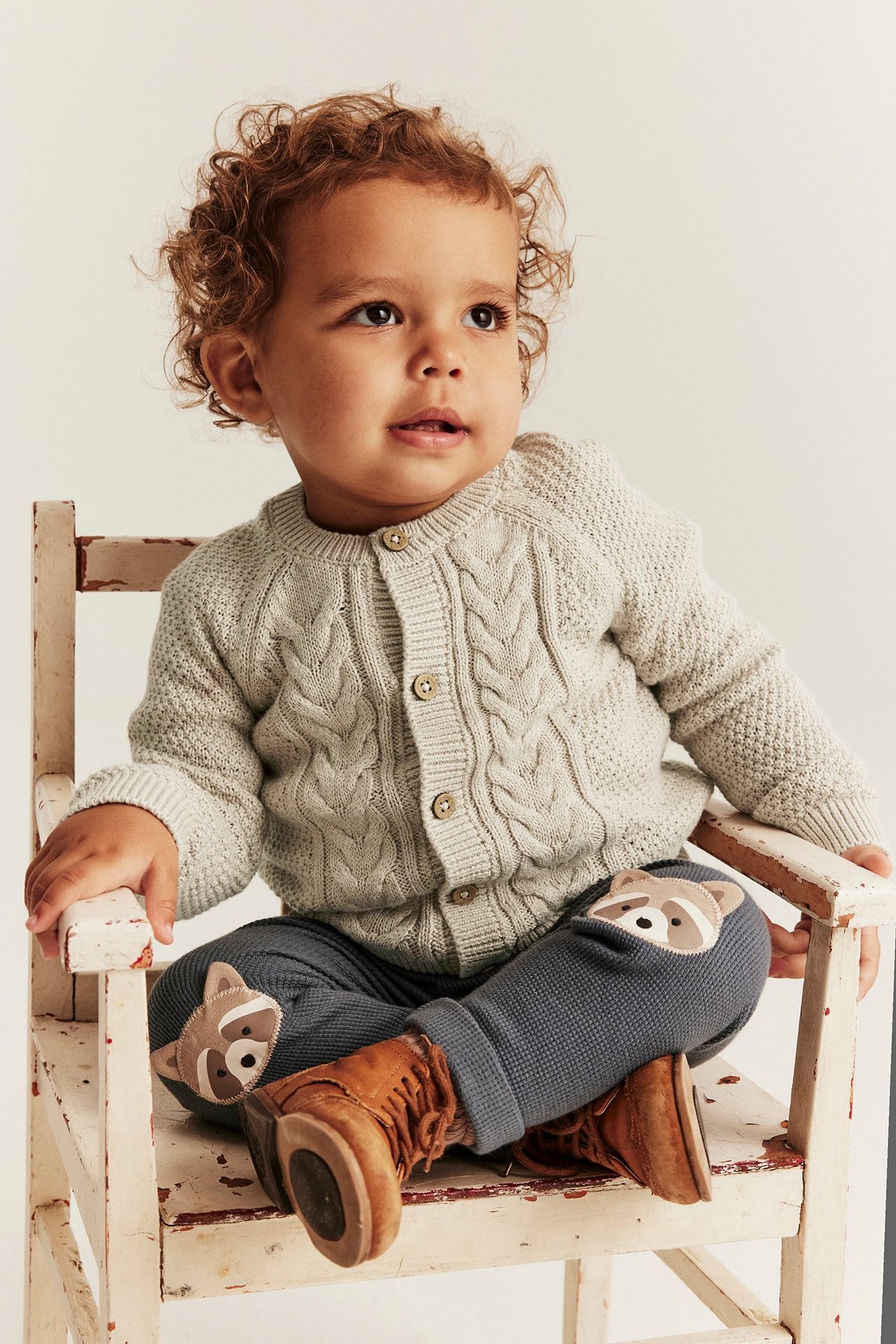 Vorderansicht Baby: beige Melange-Strickjacke mit Knöpfen, dunkelgraue Hose mit Waschbär-Patches und braune Stiefel.