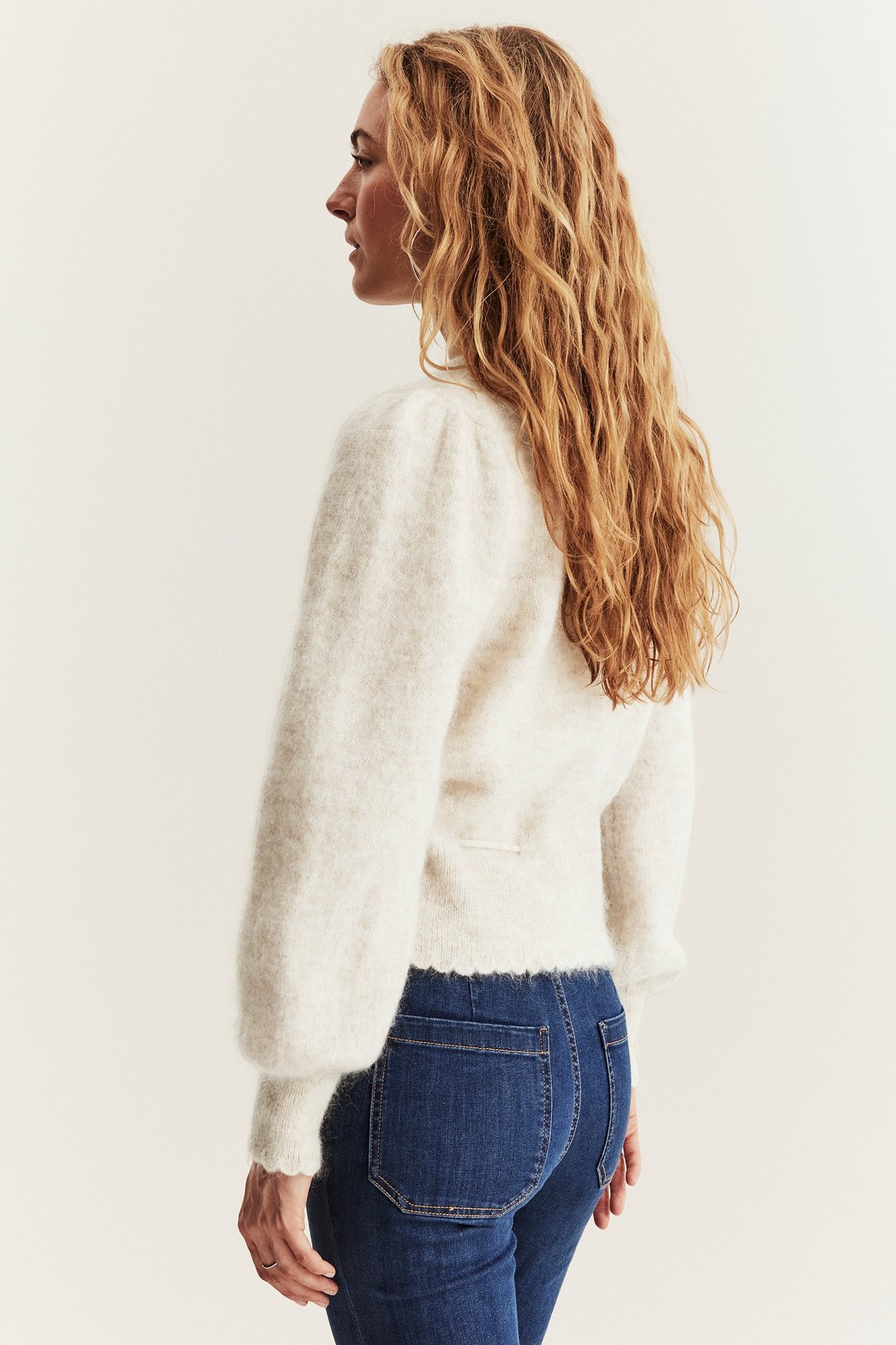 Seitenansicht einer Frau in einer hellbeigen melierten Wollmisch-Strickjacke mit Puffärmeln und dunklen Jeans.