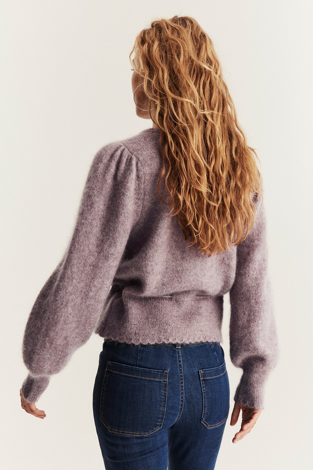 Rückenansicht einer Frau in fliederfarbener Wollmisch-Strickjacke mit Puffärmeln, Wellenkanten und dunkelblauer Jeans.