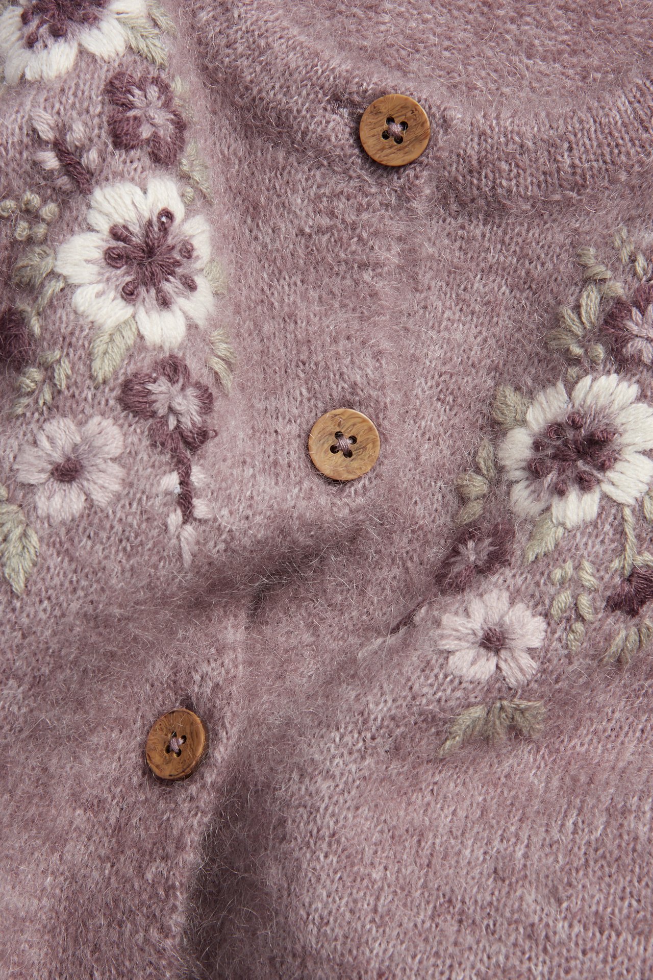 Nahaufnahme einer lila Wollmisch-Strickjacke mit weißen und lila Blumenstickereien und Holzknöpfen.