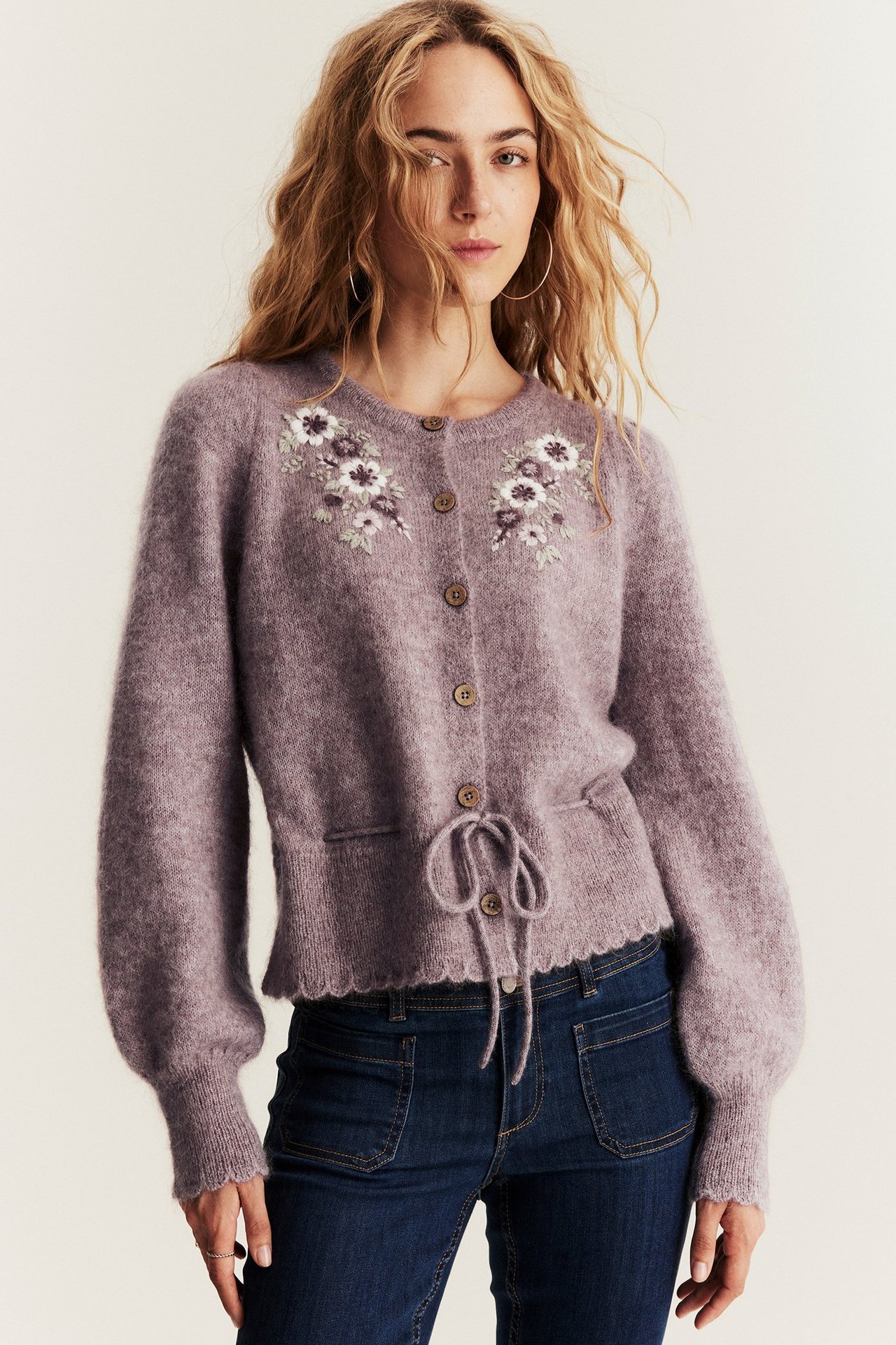 Front view of a woman wearing a lilac wool blend cardigan with embroidered flowers and dark blue jeans.
