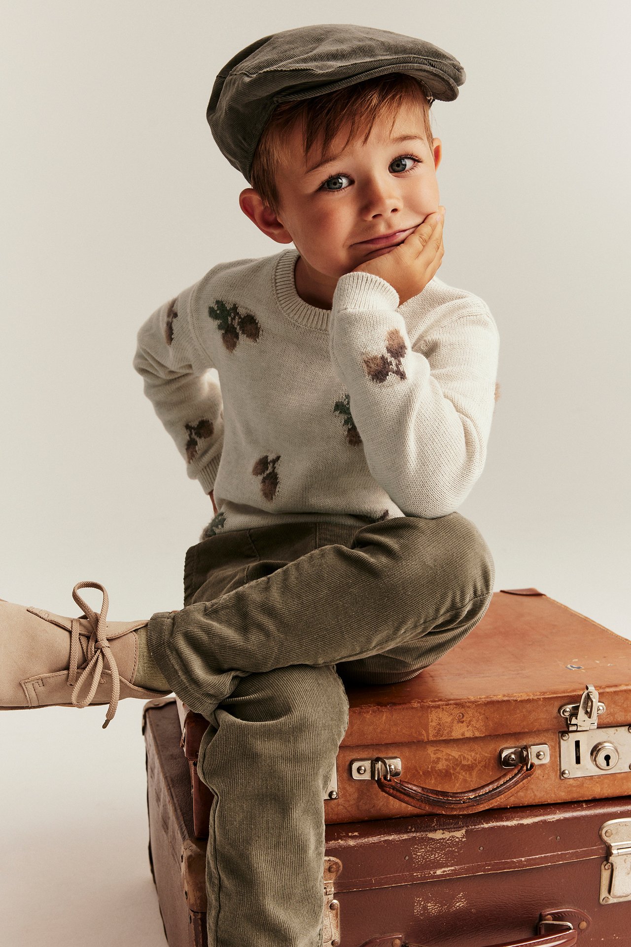 Kind in beigem Melange-Jacquard-Pullover mit Tannenzapfen, olivfarbener Hose, beigen Schuhen und grauer Mütze.