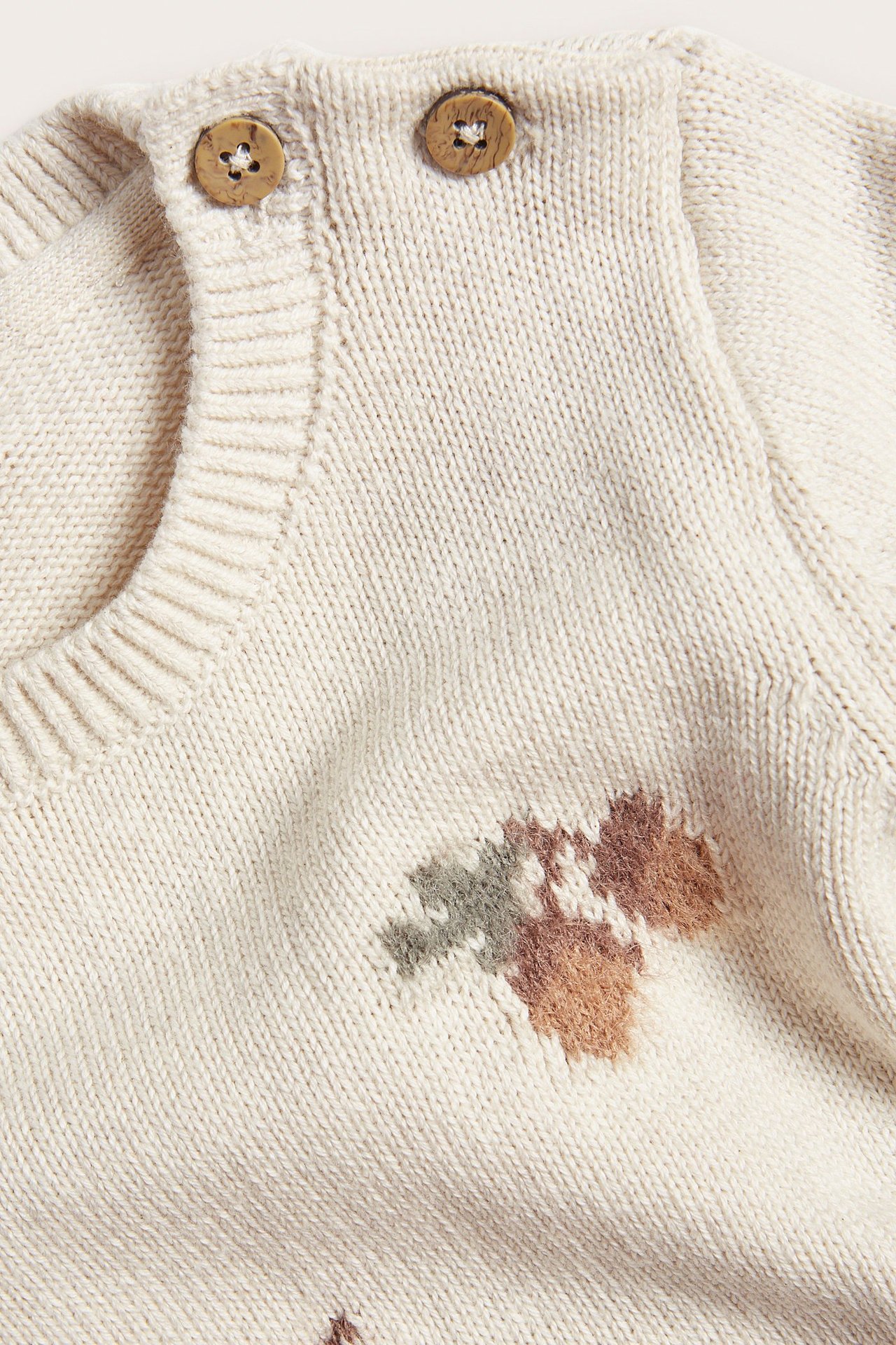 Nahaufnahme eines Baby-Pullovers in Beige Melange mit Jacquard-Blattmuster und zwei Holzknöpfen.