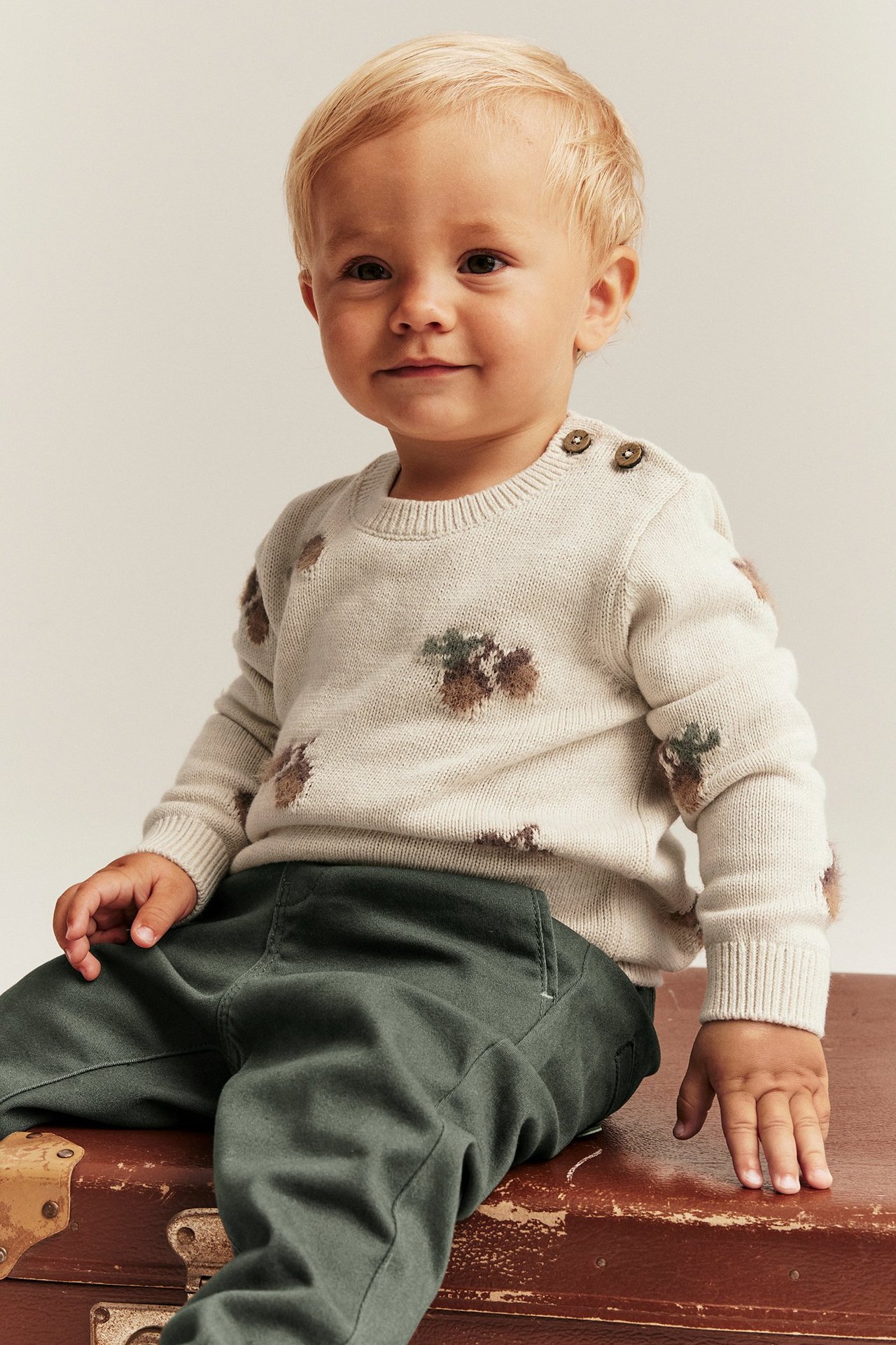 Vorderansicht: Baby trägt einen beigemelierten Jacquard-Strickpullover mit Schulterknöpfen und dunkelgrüne Hose.
