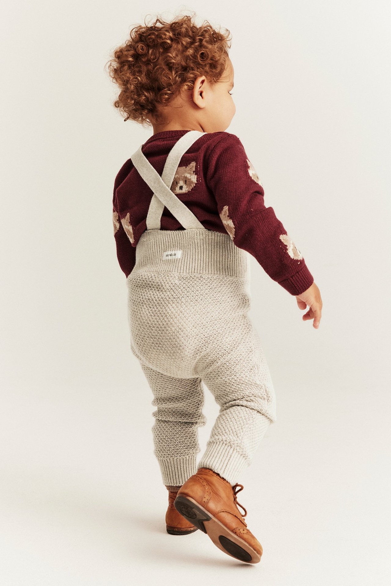 Rückenansicht: Baby trägt beige melierte Strick-Latzhose über einem weinroten Pullover mit Bärenmuster und braune Schuhe.