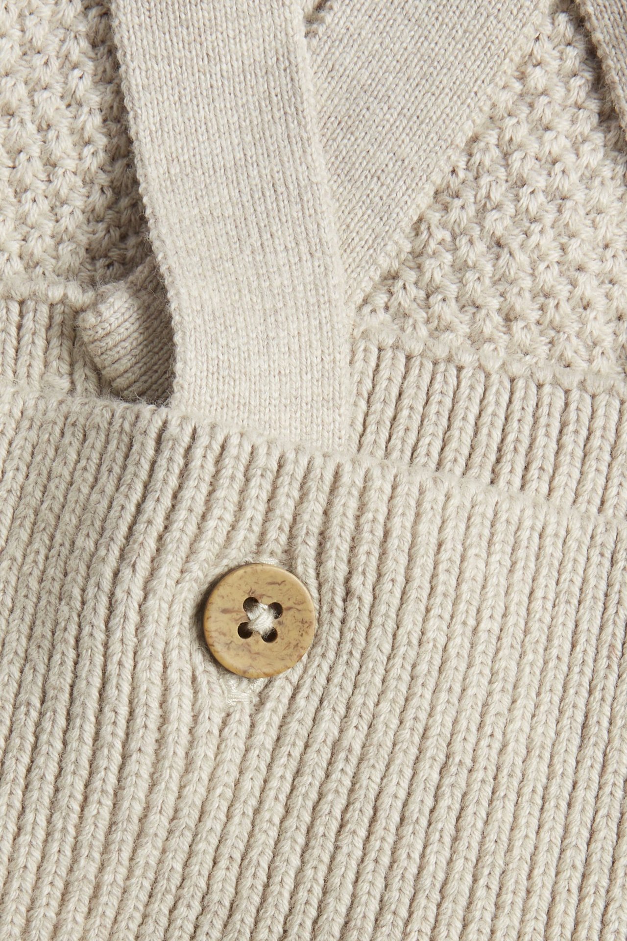 Nahaufnahme einer beige melierten, strukturgestrickten Baby-Latzhose mit verstellbarem Träger und hellbraunem Knopf.