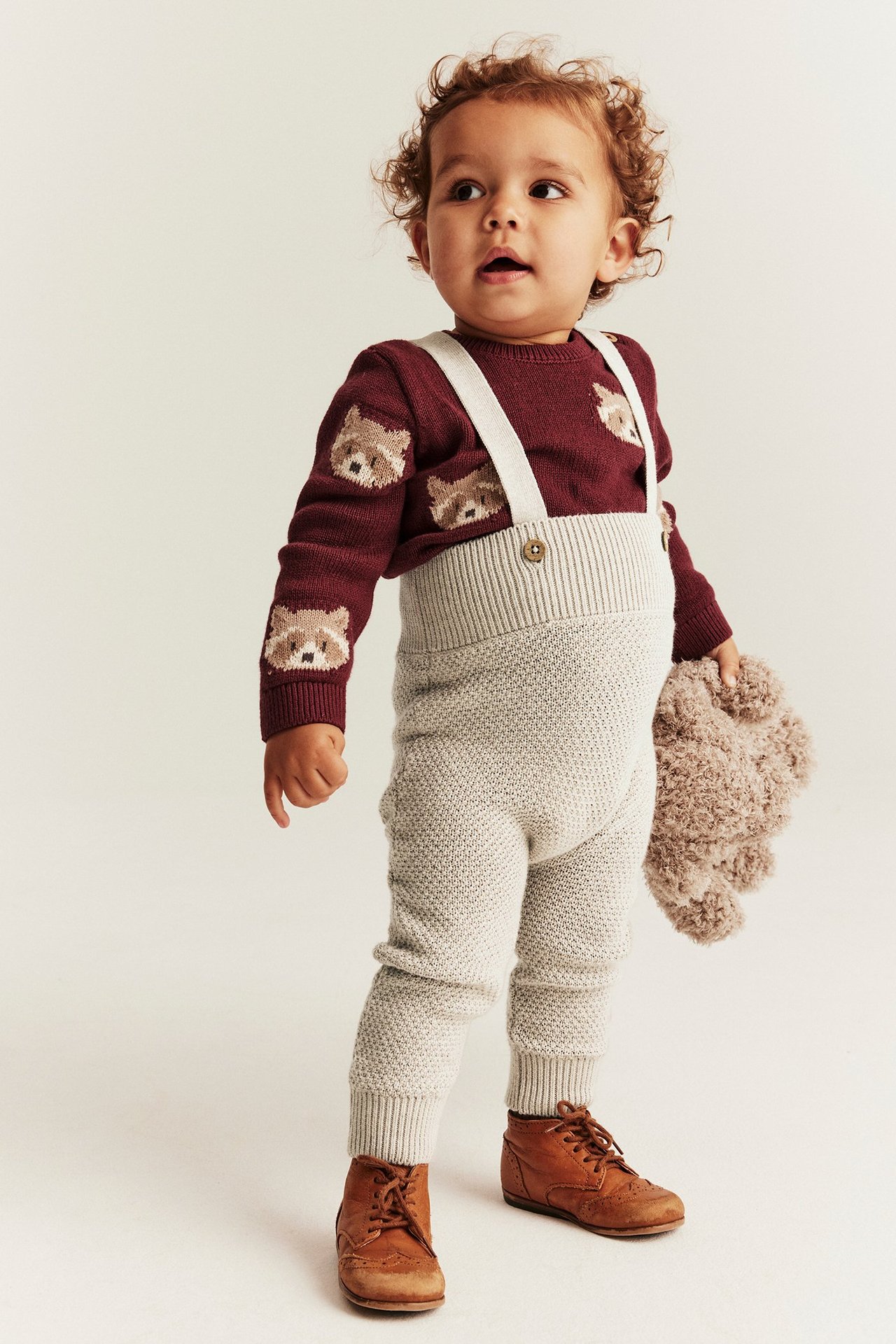 Vorderansicht eines Babys in beiger Strick-Latzhose, burgunderrotem Tiersweater, braunen Schuhen, hält einen Teddy.