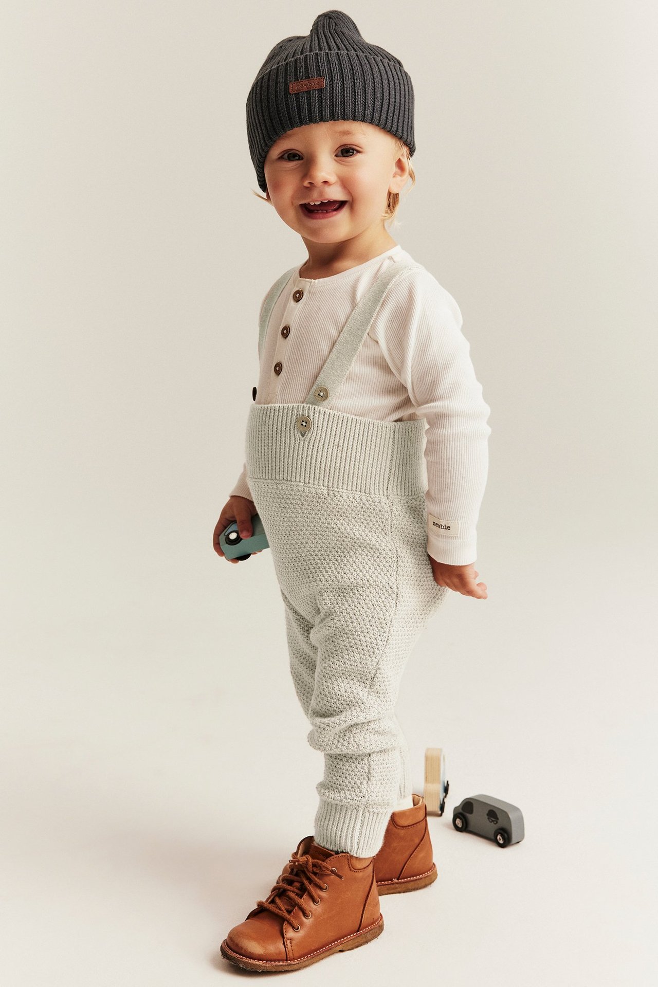 Front view: Smiling baby wears light beige knitted dungarees over white top, dark grey beanie, brown boots.