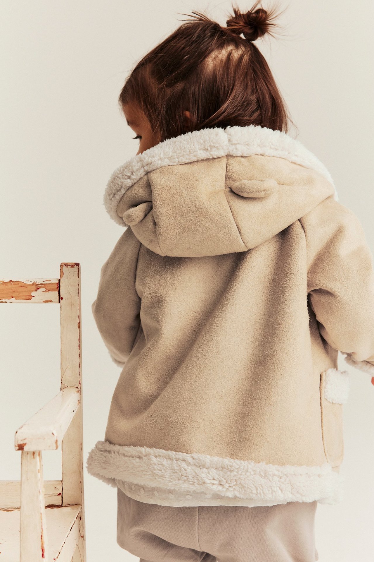 Rückenansicht: Kind in schlichter beiger Shearling-Jacke mit weißem Plüschfutter, Ohrenkapuze und hellen Hosen.