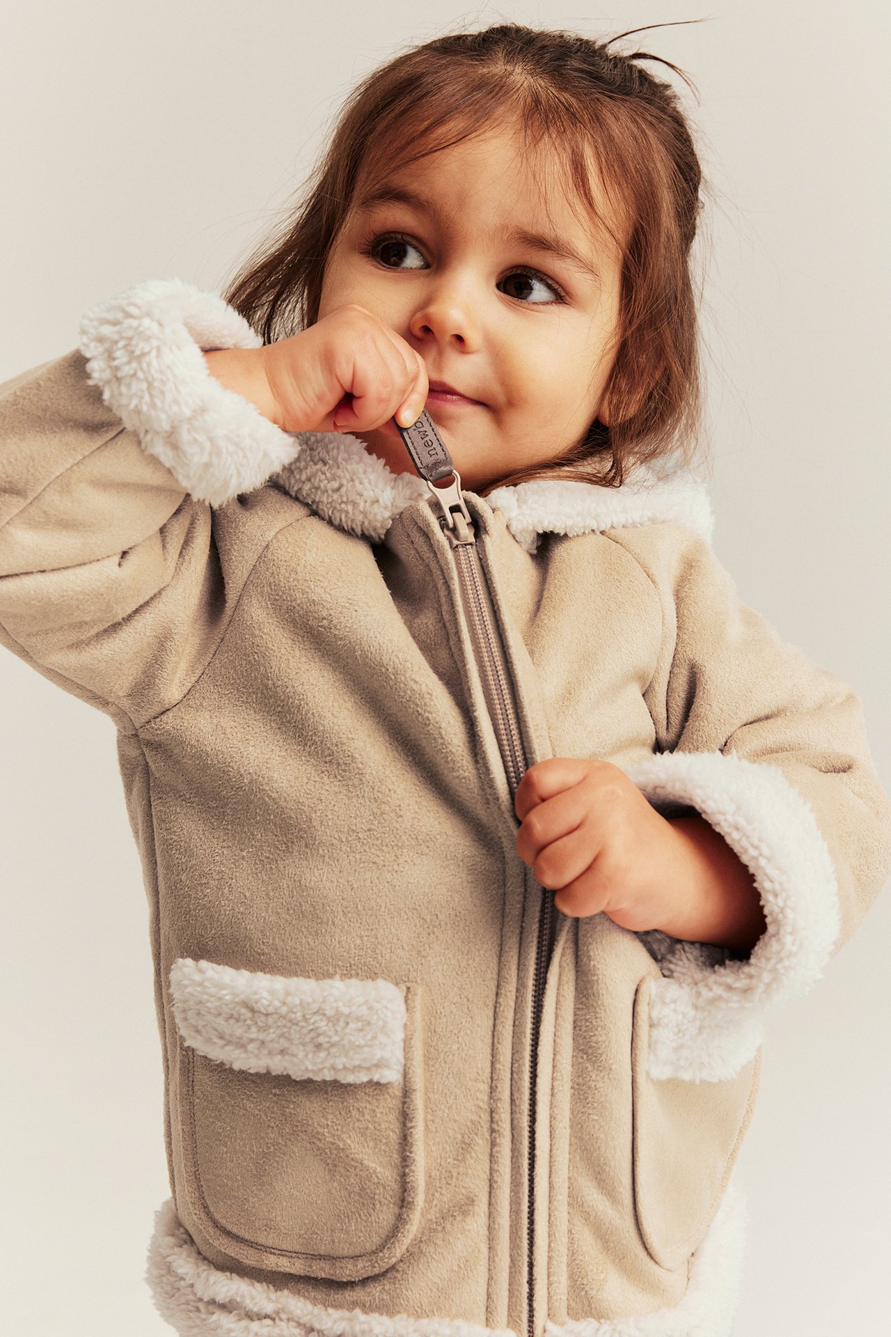 Close-up front view of a baby in a beige shearling jacket with white pile lining, holding the zipper pull.