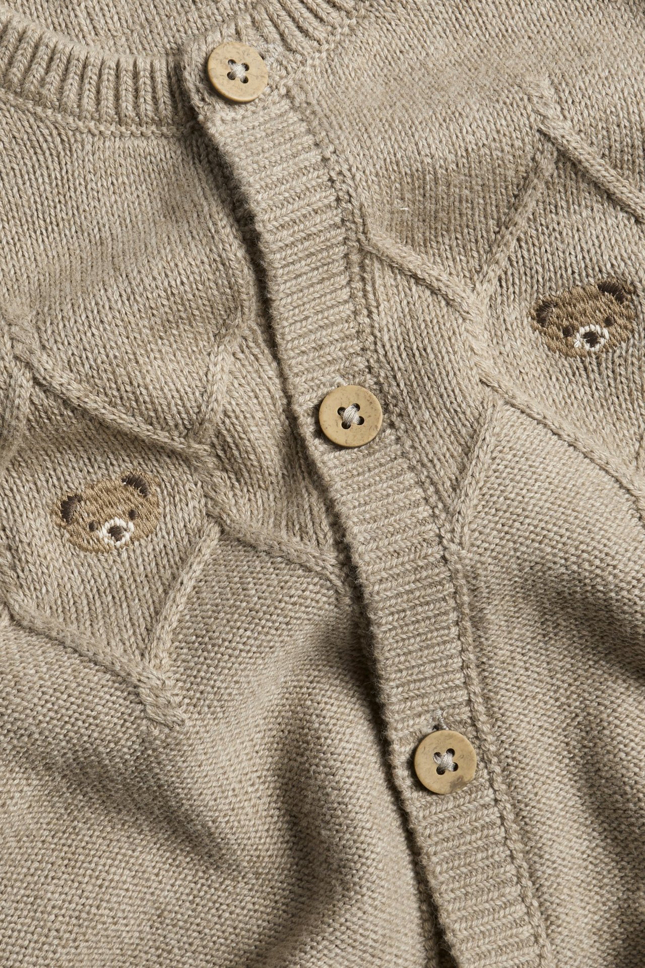 Nahaufnahme eines babybraunen melierten Strickoveralls mit Bärenstickerei und Holzknöpfen.