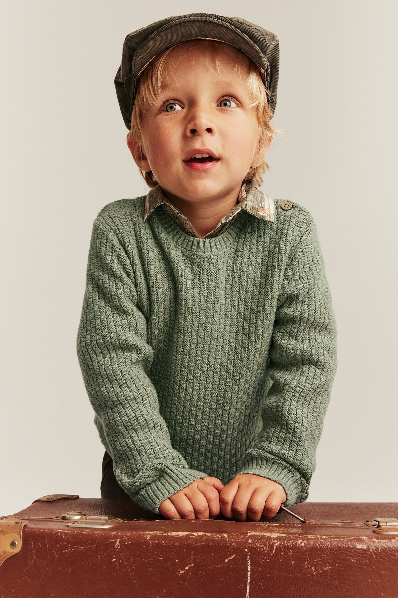 Kind in grünem Strukturstrickpullover mit Schulterknöpfen, über kariertem Hemd und dunkler Mütze.