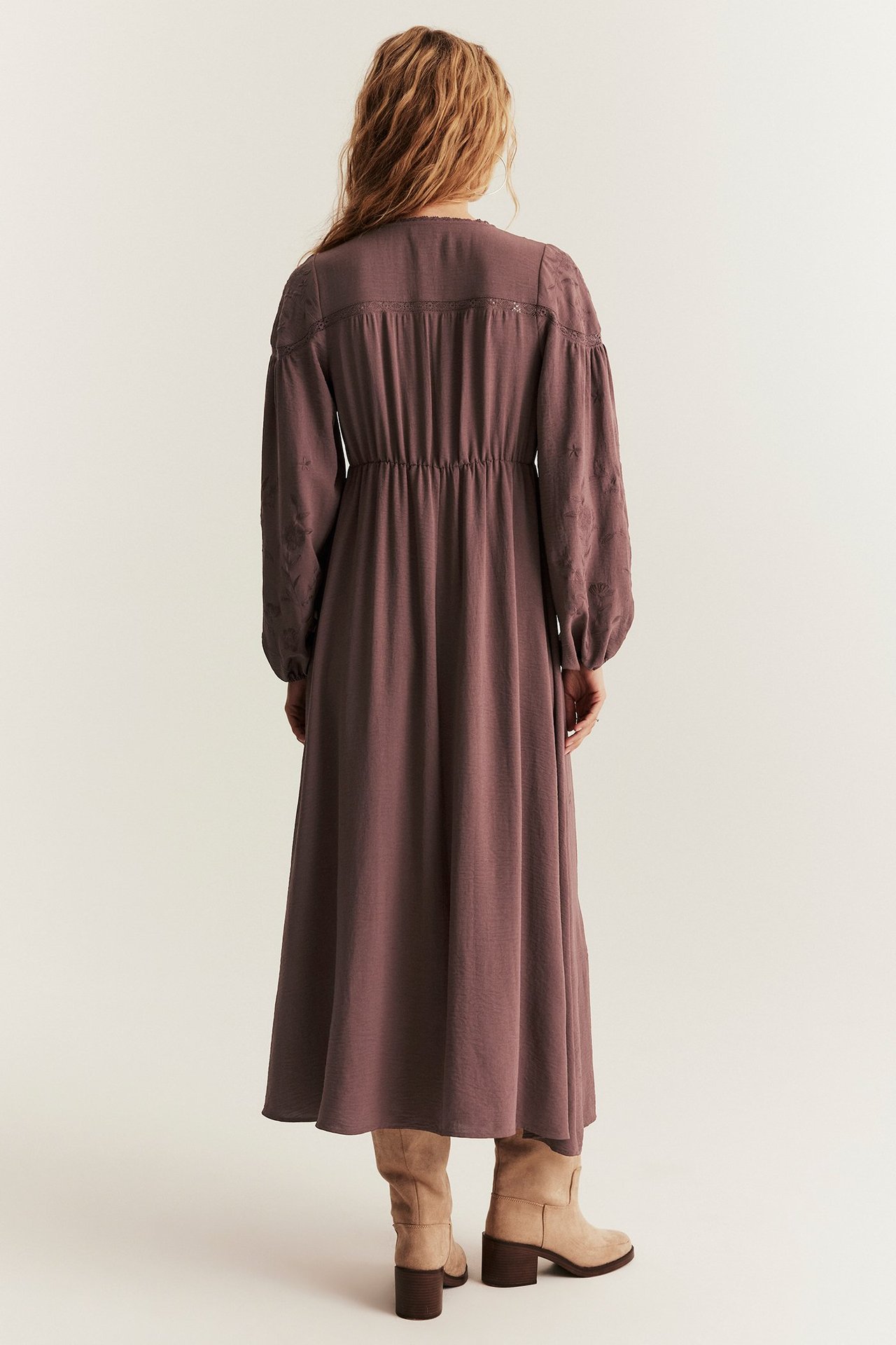 Rückenansicht: Frau in weinrotem, besticktem Langarm-Midikleid mit Gummizug und beigen Stiefeln.