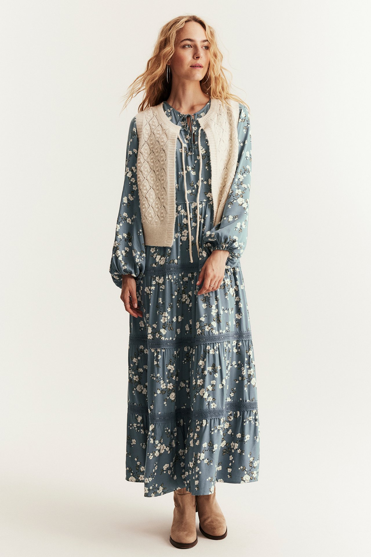 Frontansicht einer Frau in langem blauem Blumenkleid, kurzer beiger Strickweste und beigen Stiefeletten.