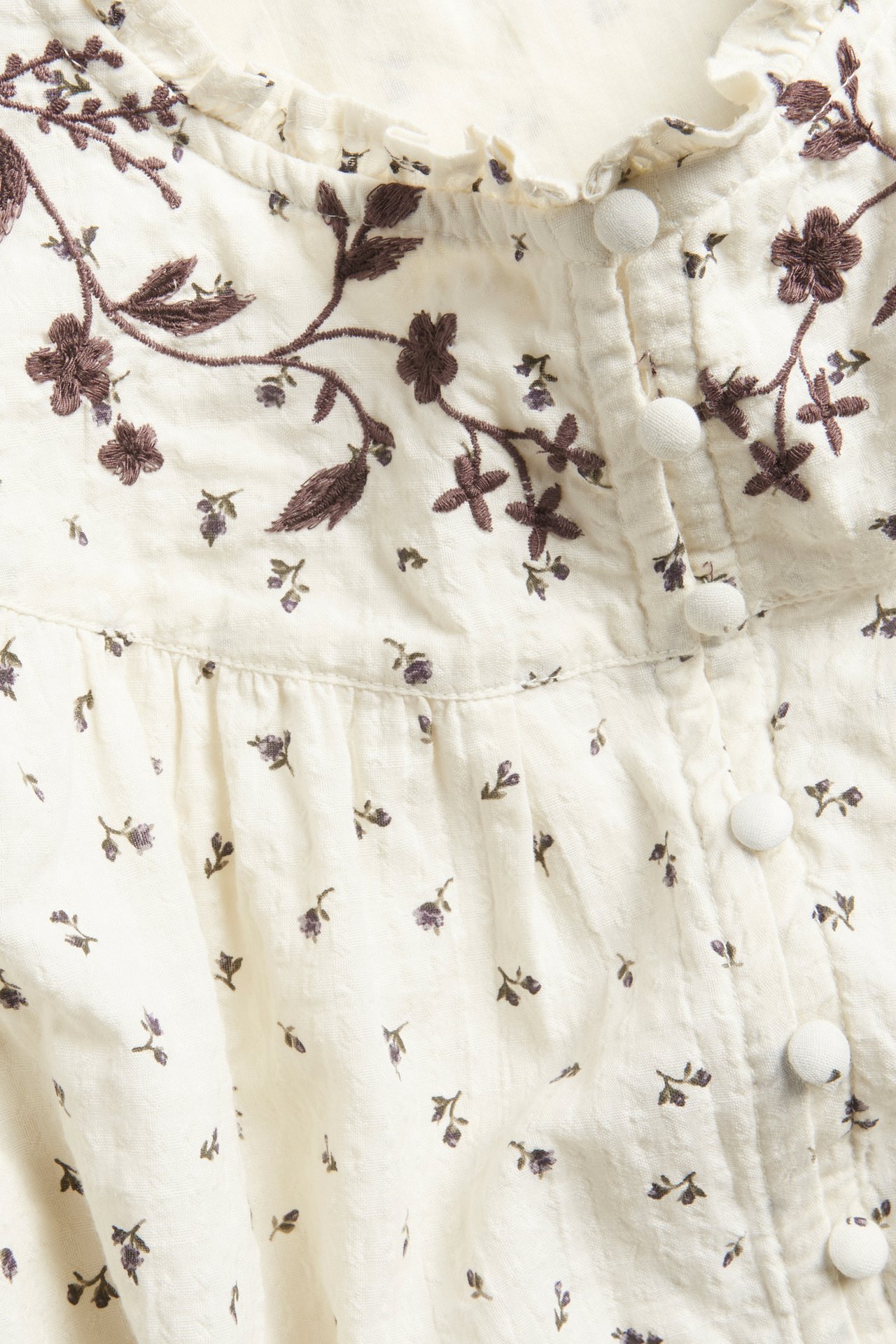 Close-up of a light beige cotton blouse with small floral print, embroidered yoke, ruffled neckline, covered buttons.