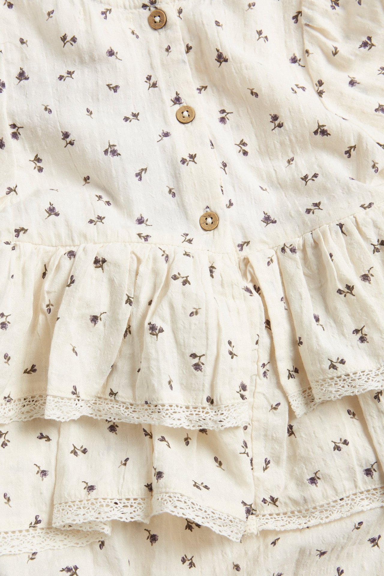 Nahaufnahme eines hellbeigen geblümten Baby-Bodys aus Webstoff mit Rüschen, Spitzenbesatz und Holzknöpfen.