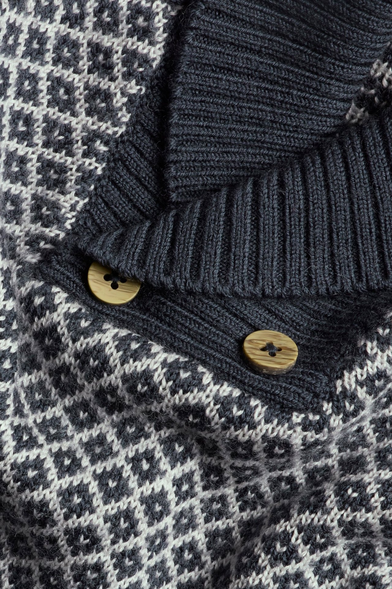 Nahaufnahme eines dunkelblauen und weiß gemusterten Wollpullovers mit Schalkragen und zwei Holzknöpfen.