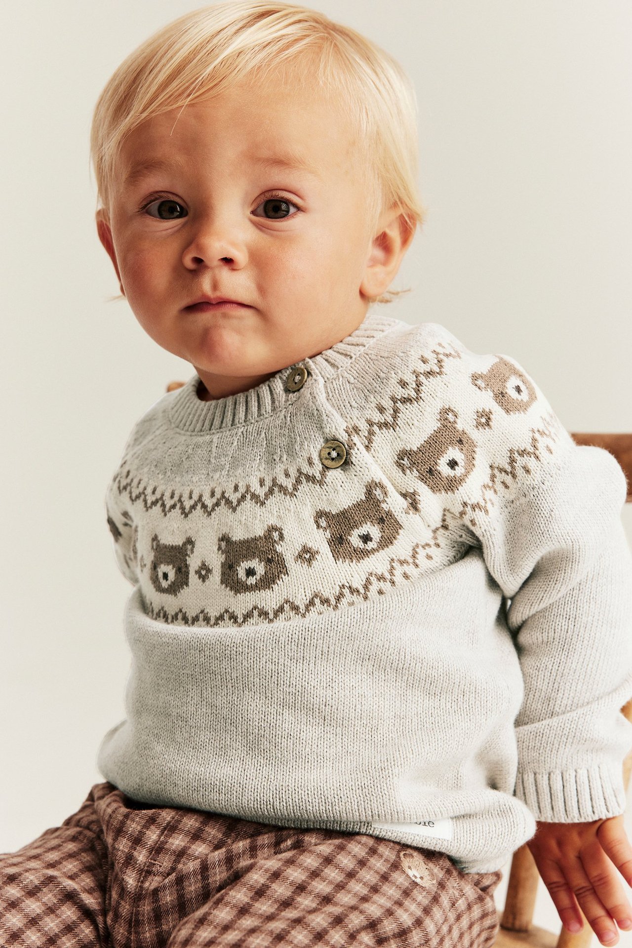 Vorderansicht eines Babys in einem beigefarbenen Jacquard-Strickpullover mit braunem Bärenmuster und karierter Hose.
