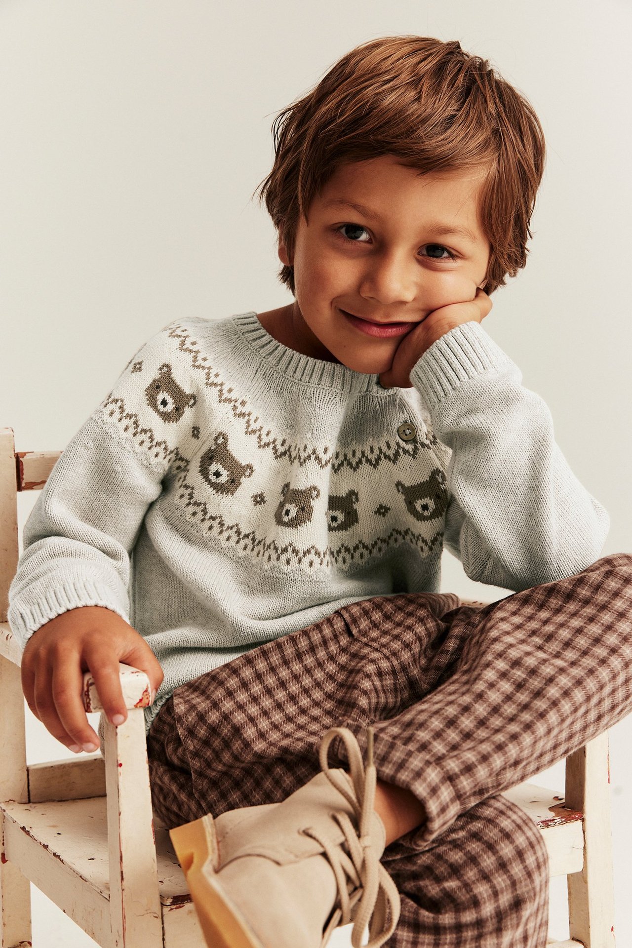 Vorderansicht lächelndes Kind in beigemelangem Jacquard-Strickpullover mit Bärenmuster und brauner Karohose.