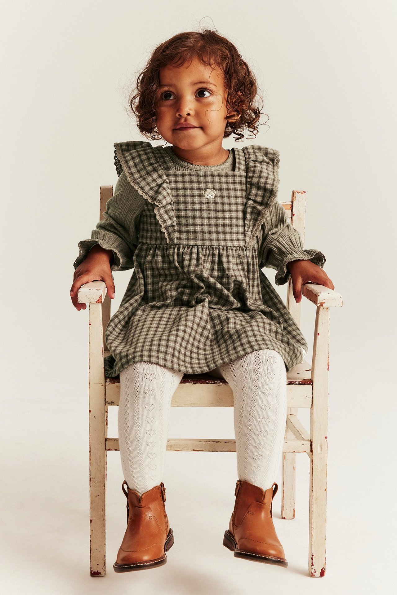 Vorderansicht: Kind trägt braunes kariertes Latzkleid mit Teddy, grünes Oberteil, weiße Strumpfhose, braune Stiefel.