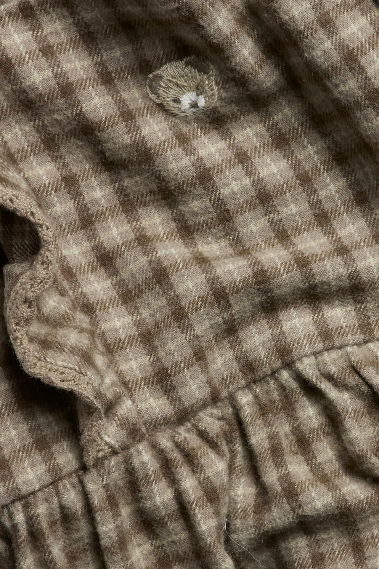 Nahaufnahme eines braun karierten Baumwoll-Latzkleides mit gesticktem Teddybär und spitzenbesetzten Rüschen.