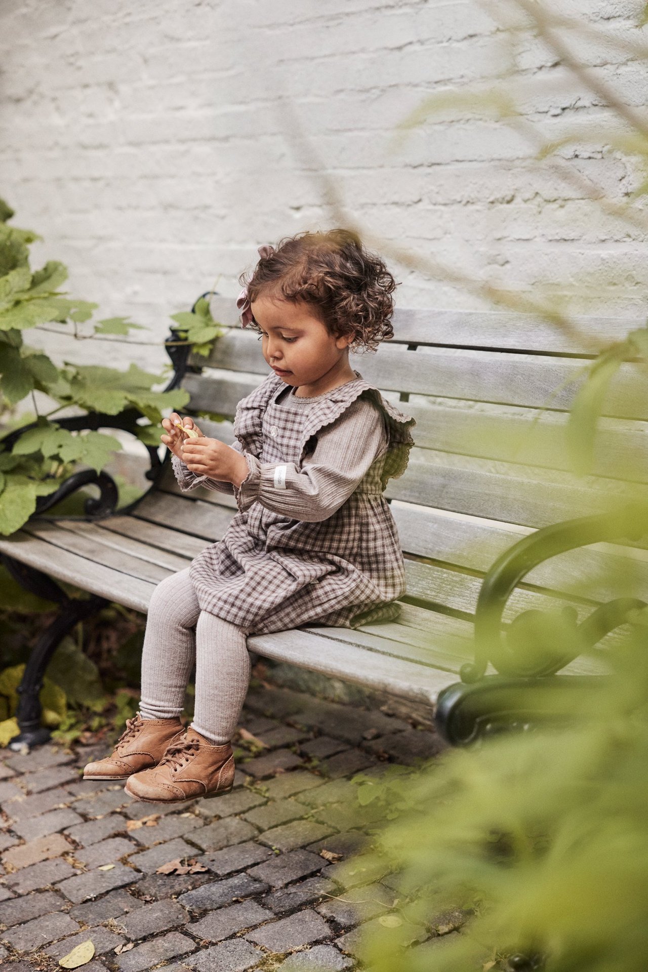 Seitenansicht: Kind in braun kariertem Rüschen-Latzkleid, Rippoberteil, beiger Strumpfhose, braunen Schnürstiefeln.