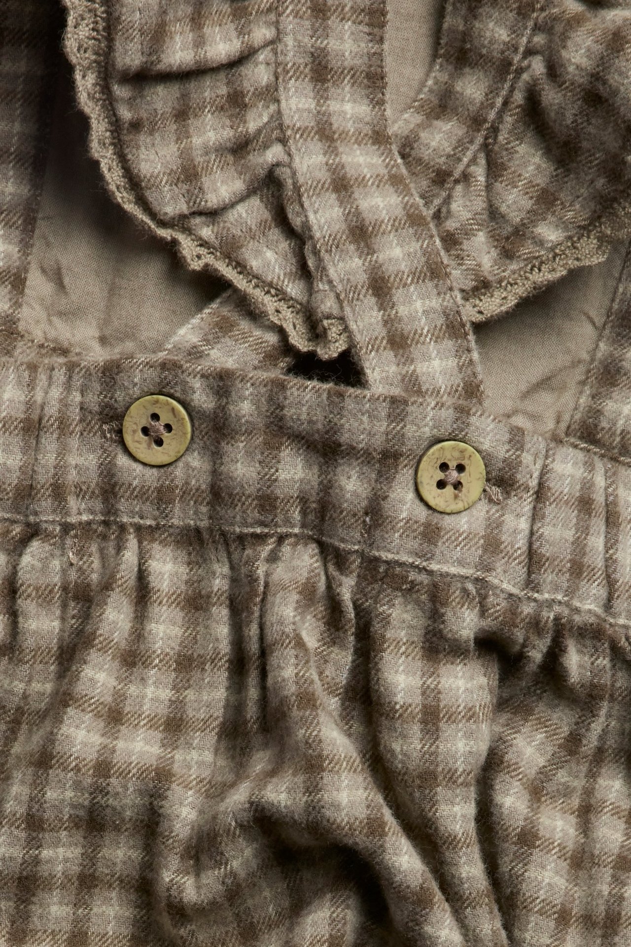 Close-up of a brown checked pinafore dress with lace-edged ruffled straps and two buttons.