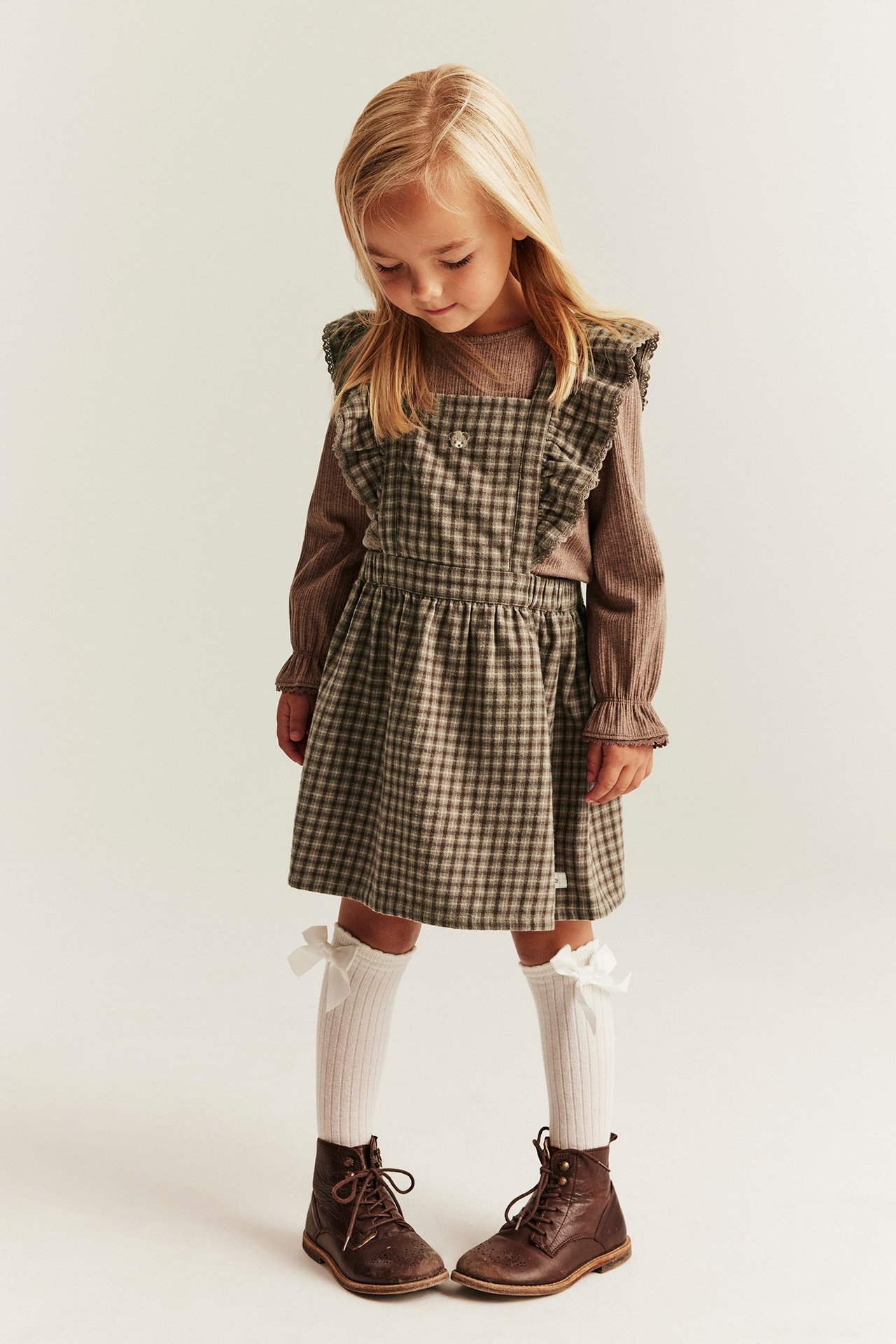 Front view: Kid in brown checked pinafore dress with ruffled straps, brown top, white socks, brown boots.