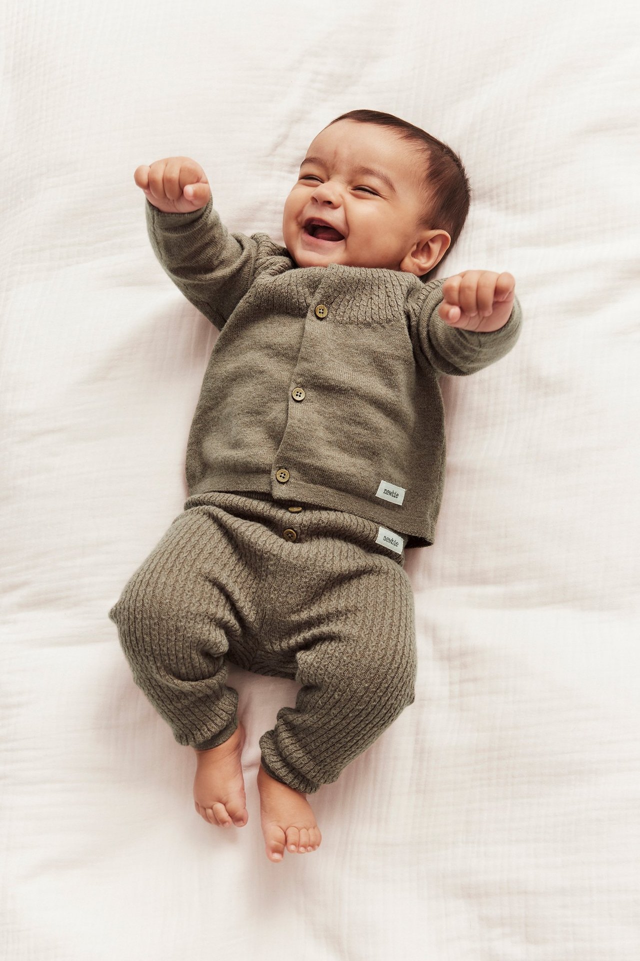 Top-down view of a laughing baby wearing brown melange structured knit leggings with decorative buttons and a matching cardigan.