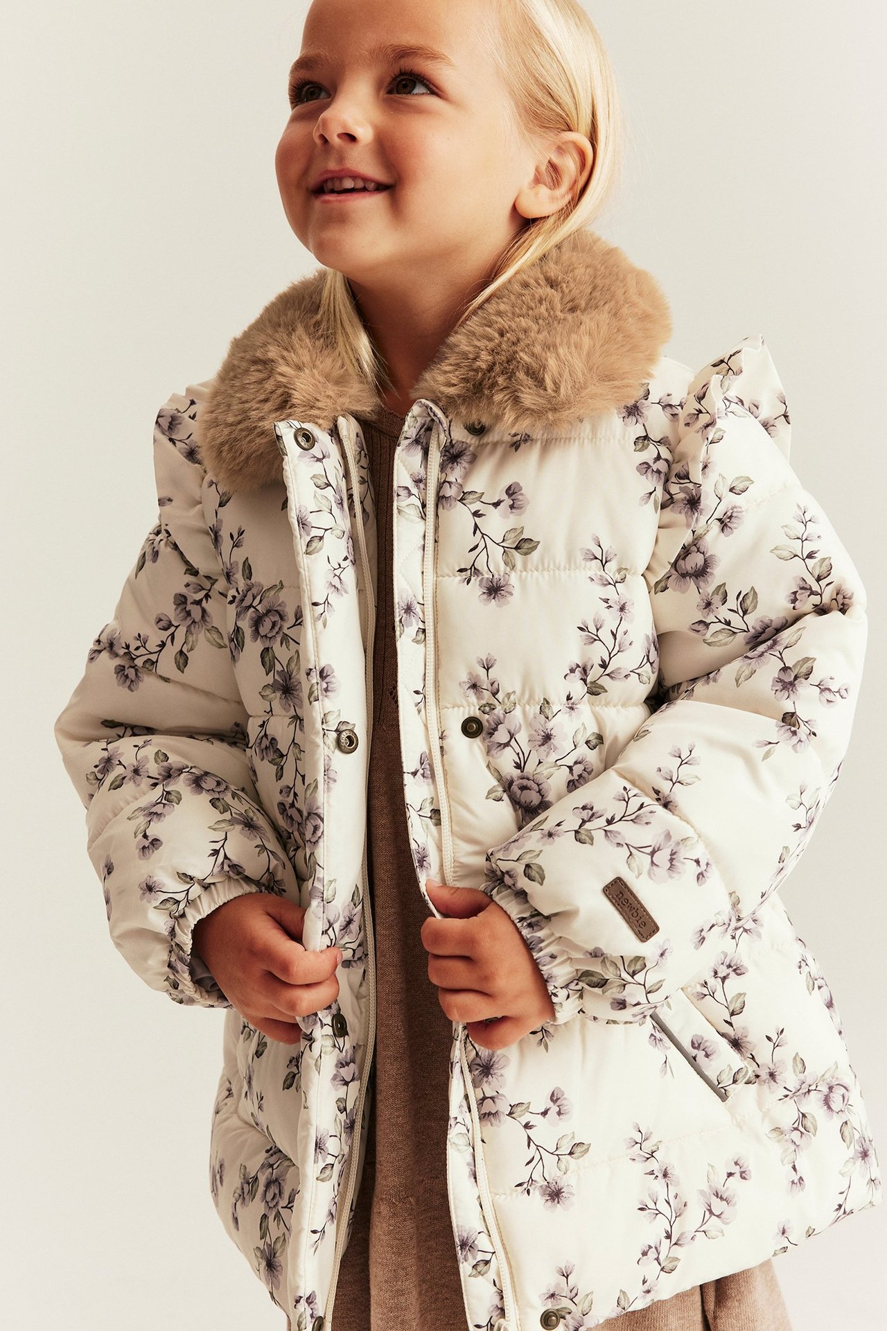 Vorderansicht: Kind in hellbeiger geblümter Steppjacke mit braunem Kunstfellkragen und Reißverschluss.