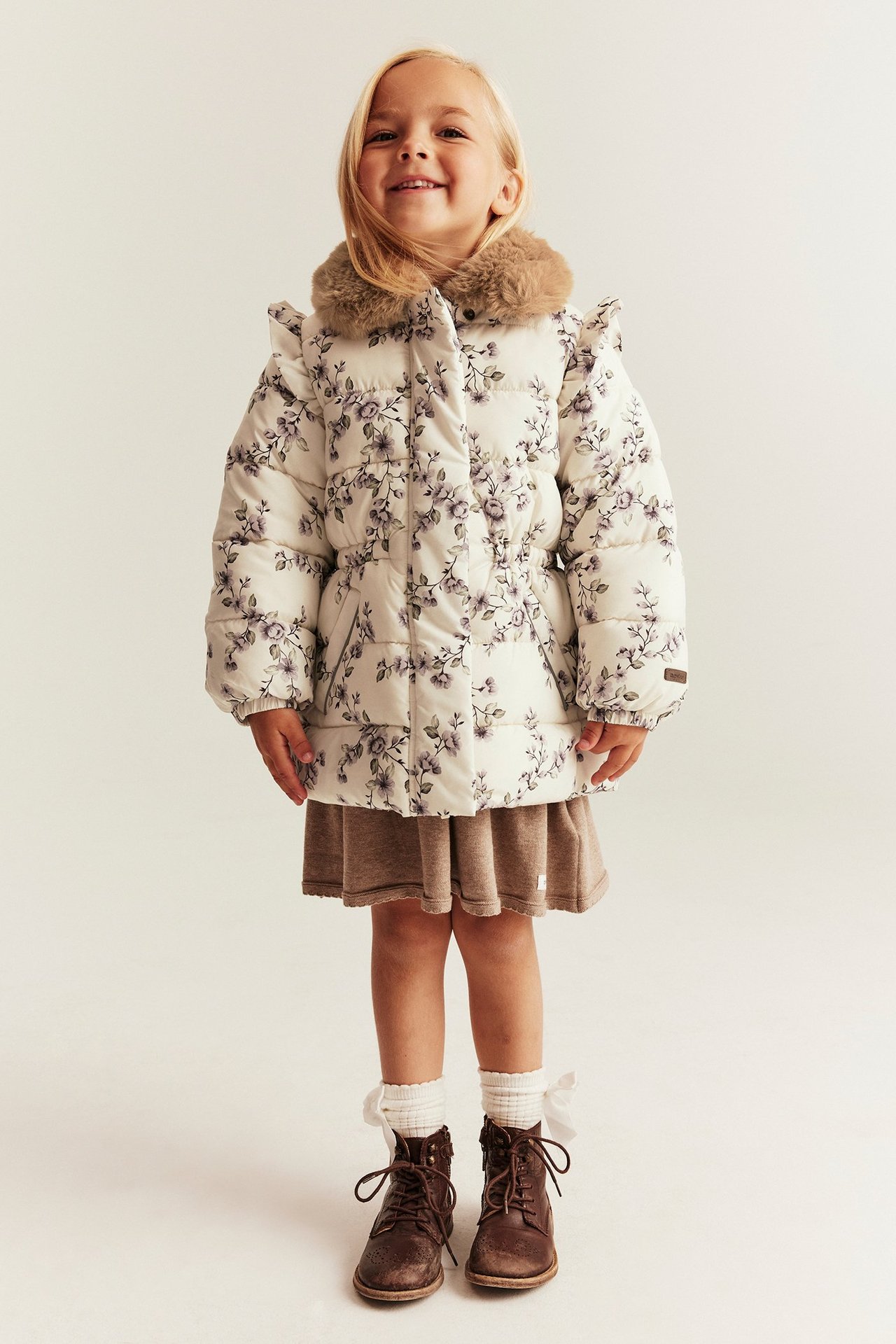 Vorderansicht: Kind trägt eine geblümte hellbeige Jacke, Kunstfellkragen, braunes Kleid, weiße Socken, braune Stiefel.